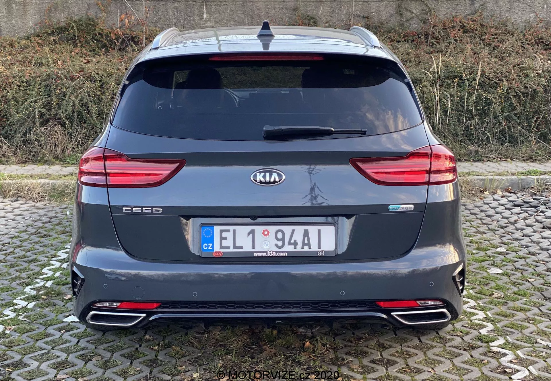 Pohľad zozadu na Kia Ceed 2018 zobrazujúci celú zadnú časť s logami značky a modelu, modernými LED zadnými svetlami, zadným oknom, dvojitými výfukovými koncovkami, zadným nárazníkom s odrazkami, strešným spojlerom, anténou a zadnými parkovacími senzormi. Obraz je jasný, urobený vo výške očí s prirodzeným osvetlením za podmienok zamračenej oblohy.