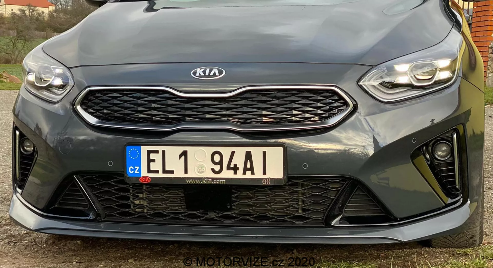 Tento obrázok zobrazuje prednú časť vozidla Kia Ceed 2018 nasnímanú z nízkeho uhla. Zdôrazňuje elegantné, horizontálne orientované LED svetlomety, výraznú mriežku so vzorom včelieho plástu so stredovo umiestneným emblémom Kia, európsku poznávaciu značku z Českej republiky a športový predný nárazník s veľkými otvormi. Bližší pohľad zvýrazňuje parkovacie senzory pod mriežkou. Športový dizajn naznačuje vyšší výkonový variant, aj keď presné špecifikácie nie sú jasné. Obraz s vysokým rozlíšením zachytáva dizajnové detaily a kontúry a je mierne posunutý doľava.