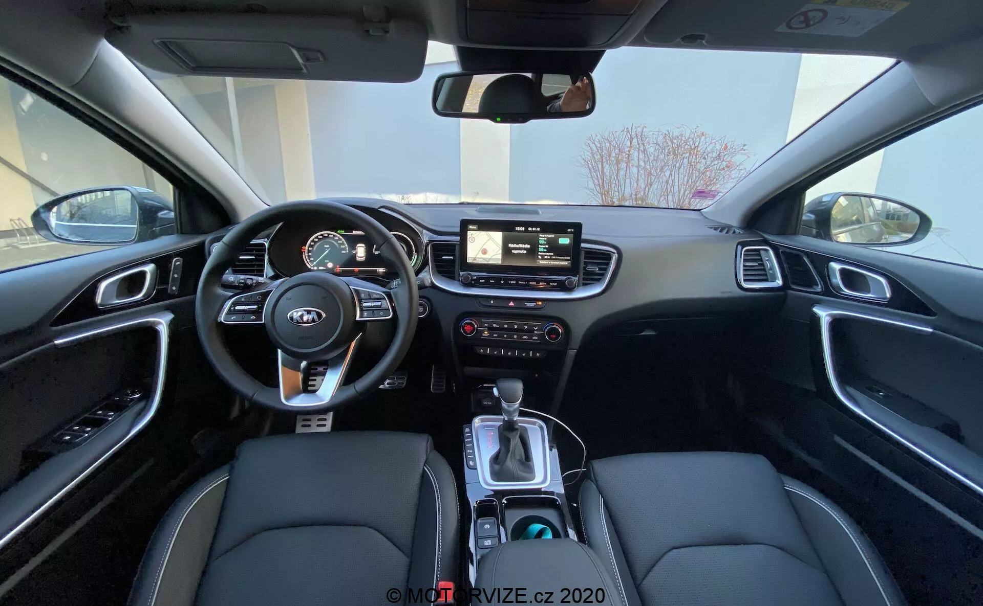 Širokouhlý pohľad na interiér Kia Ceed 2018 zo zadného sedadla, zobrazujúci oblasť vodiča vrátane volantu s ovládacími prvkami, prístrojovej dosky s digitálnymi a analógovými ukazovateľmi, centrálneho infotainment systému s dotykovou obrazovkou, manuálnymi ovládacími prvkami pre infotainment a klimatizáciu, a stredovej konzoly s radiacou pákou, voličom jazdného režimu, držiakmi na nápoje a čierneho interiéru s látkovým poťahom zo syntetickej kože s bielym prešívaním a hliníkovým obložením.