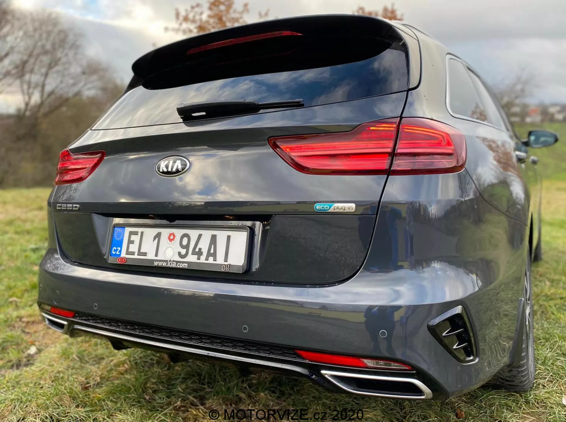 Zadný pohľad na tmavosivý model Kia Ceed Proceed 2018 so zaujímavými prvkami, ako sú horizontálne orientované zadné LED svetlá, strešný zadný spojler, dvojité chrómované výfuky, zadný nárazník v štýle difúzora a centrálne znaky vrátane 'Kia' a 'GT Line'. Kamera na cúvanie nad centrálne umiestnenou ŠPZ naznačuje parkovaciu asistenciu, zatiaľ čo plytká hĺbka ostrosti zdôrazňuje značku a identifikátory modelu auta na pozadí s trávou.