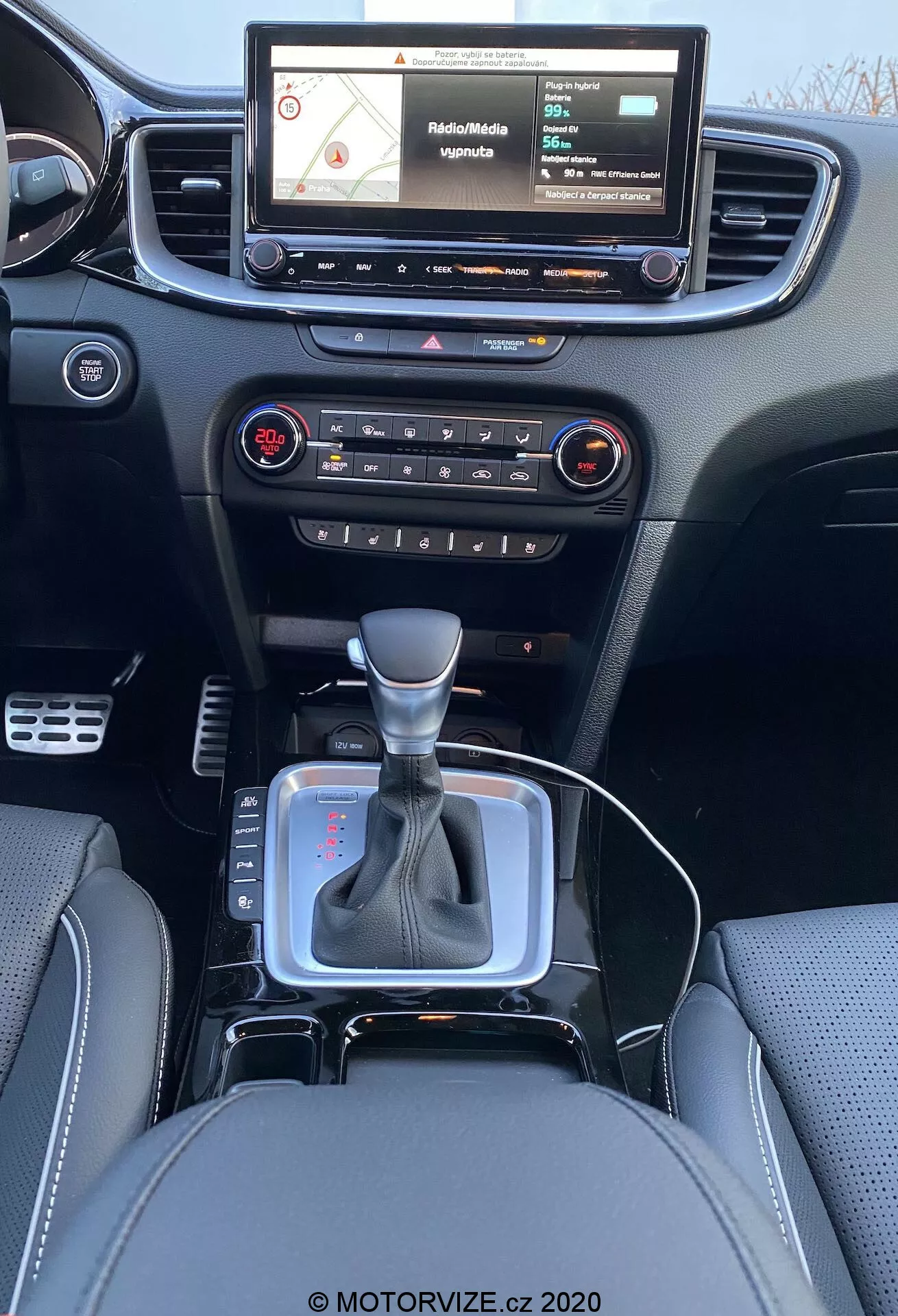 Interiérový pohľad na Kia Ceed 2018 z pohľadu zadného sedadla, zobrazujúci modernú prístrojovú dosku s dotykovým infotainment systémom ukazujúcim navigačnú mapu, fyzickými tlačidlami a ovládačmi pre kontrolu audia a klimatizácie, centrálnym výduchom vzduchu, radiacou pákou v neutrálnej polohe so strieborným akcentom a čiernym koženým poťahom, malý úložný priestor pri radiacej páke pravdepodobne pre 12V zásuvku alebo USB porty, kožené sedadlá s detailným prešívaním.