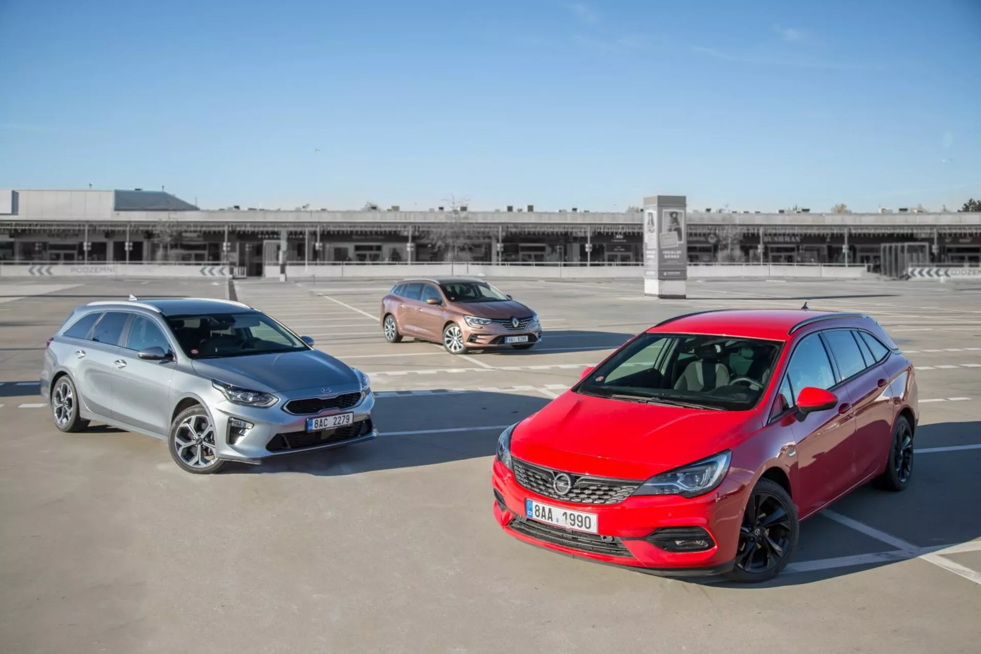 Tri modely Kia Ceed z roku 2009 vrátane červeného hatchbacku, zlatého crossoveru a strieborného kombi zaparkované na strešnom parkovisku, odfotené z vyvýšenej pozície s výhľadom na dizajn prednej časti, bočné profily a dĺžku vozidiel