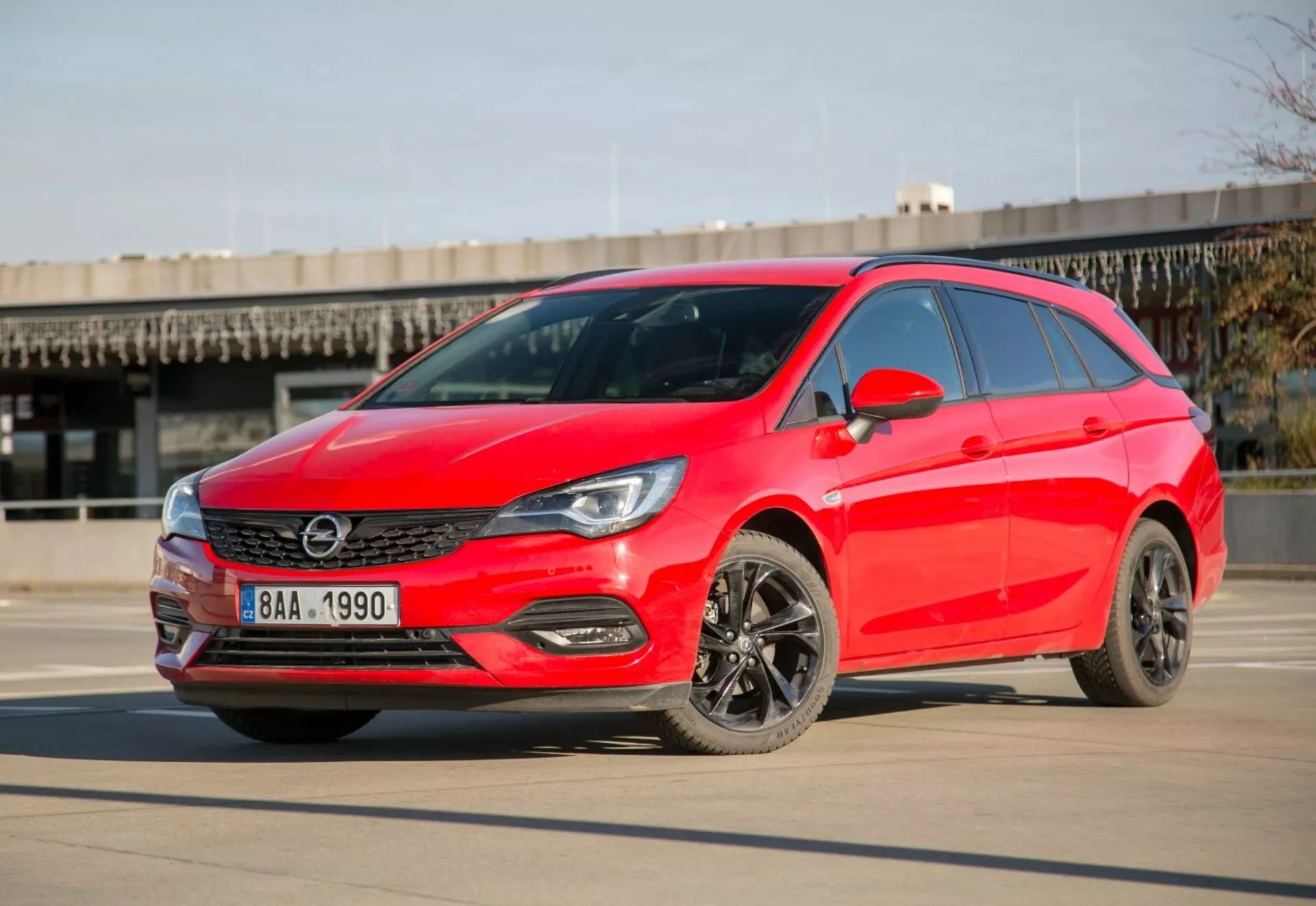 Jasne červený hatchback Kia Ceed z roku 2009 zaparkovaný na hladkom betóne pod jasnou oblohou, predvádzajúci LED svetlomety, elegantnú mriežku, tmavé zliatinové kolesá a moderný aerodynamický dizajn z uhla tri štvrtiny spredu počas dňa.