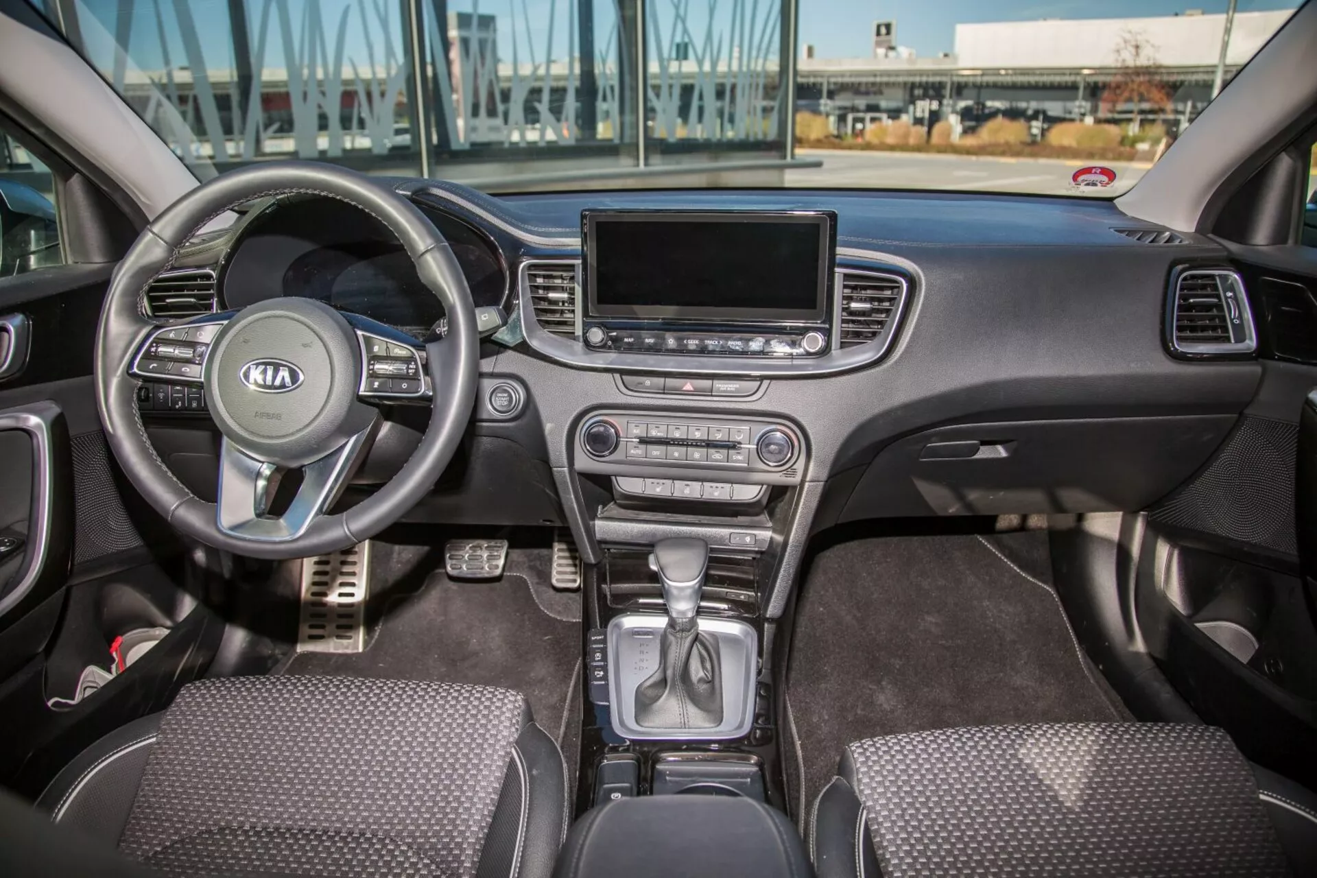 Interiér Kia Cee'd z roku 2009 s výhľadom na celú prednú kabinu vrátane multifunkčného volantu s logom Kia, časti prístrojovej dosky za volantom, radenie páky v parkovacej pozícii na stredovej konzole, tlačidiel klímy, obrazovky infotainmentu, sedadiel s povrchom z látky a ovládania dverí vodiča a spolujazdca pri prirodzenom dennom svetle