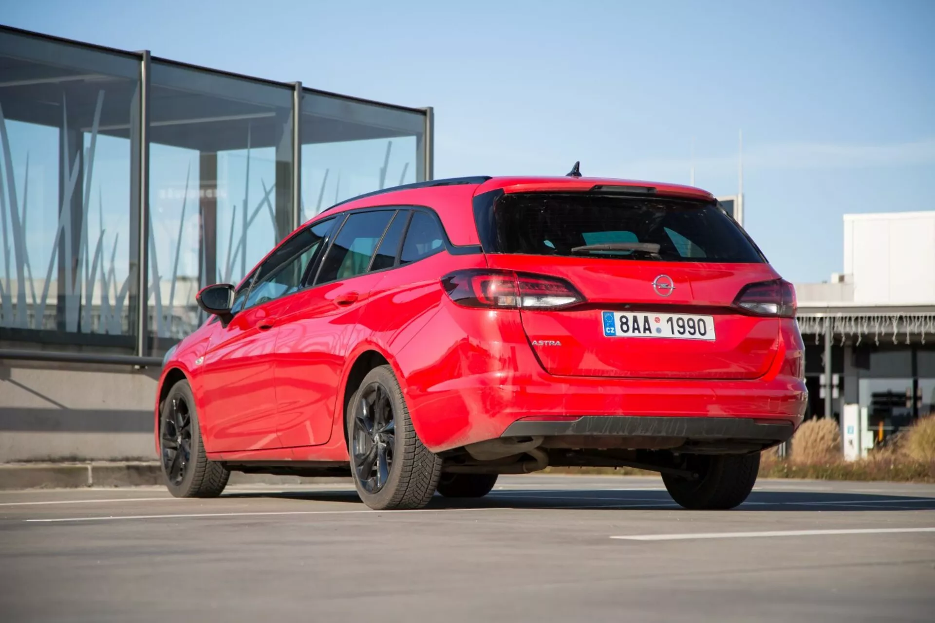 Červený hatchback Kia Ceed 2009 ukazuje svoj športový zadný trojštvrťový pohľad na betóne s pozadím moderného budovy so sklenenou fasádou, predvádza výrazný dizajn zadných svetiel, spoiler na streche, anténu žraločej plutvy, zliatinové ráfiky a možné parkovacie senzory.