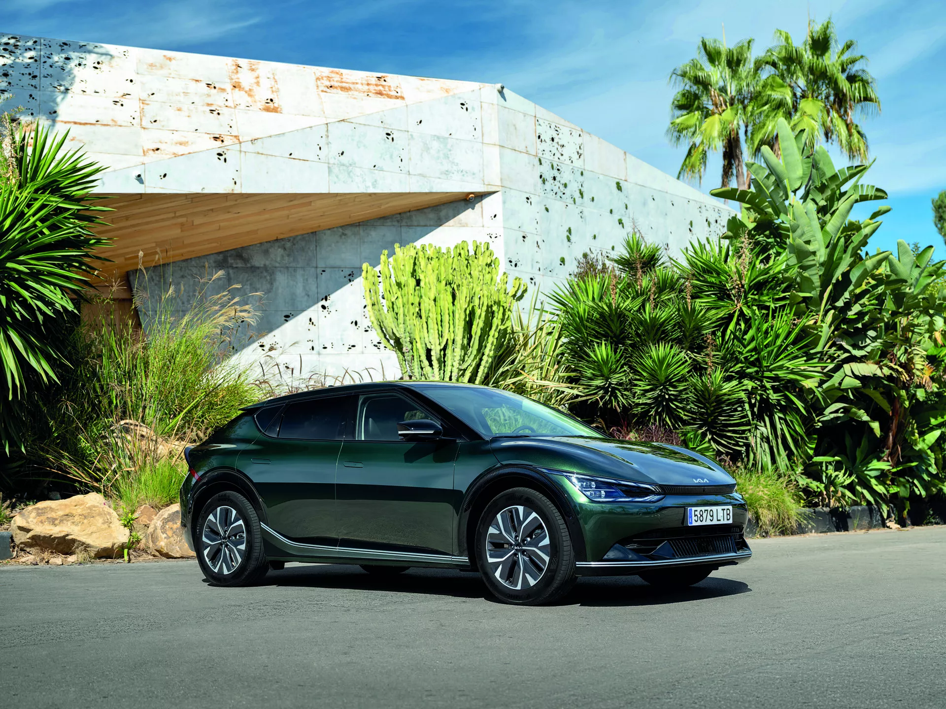 Kia EV6 z roku 2021 zachytená z nízkeho uhla zdôrazňujúca jej elegantný dizajn, športový postoj s trojštvrťovým pohľadom zo prednej strany ukazujúci LED svetlomety, aerodynamické telo, zliatinové kolesá, integrované smerové signály na bočných zrkadlách, zaparkovaná vonku pod jasným denným svetlom s modernou budovou a bujnou zeleňou na pozadí
