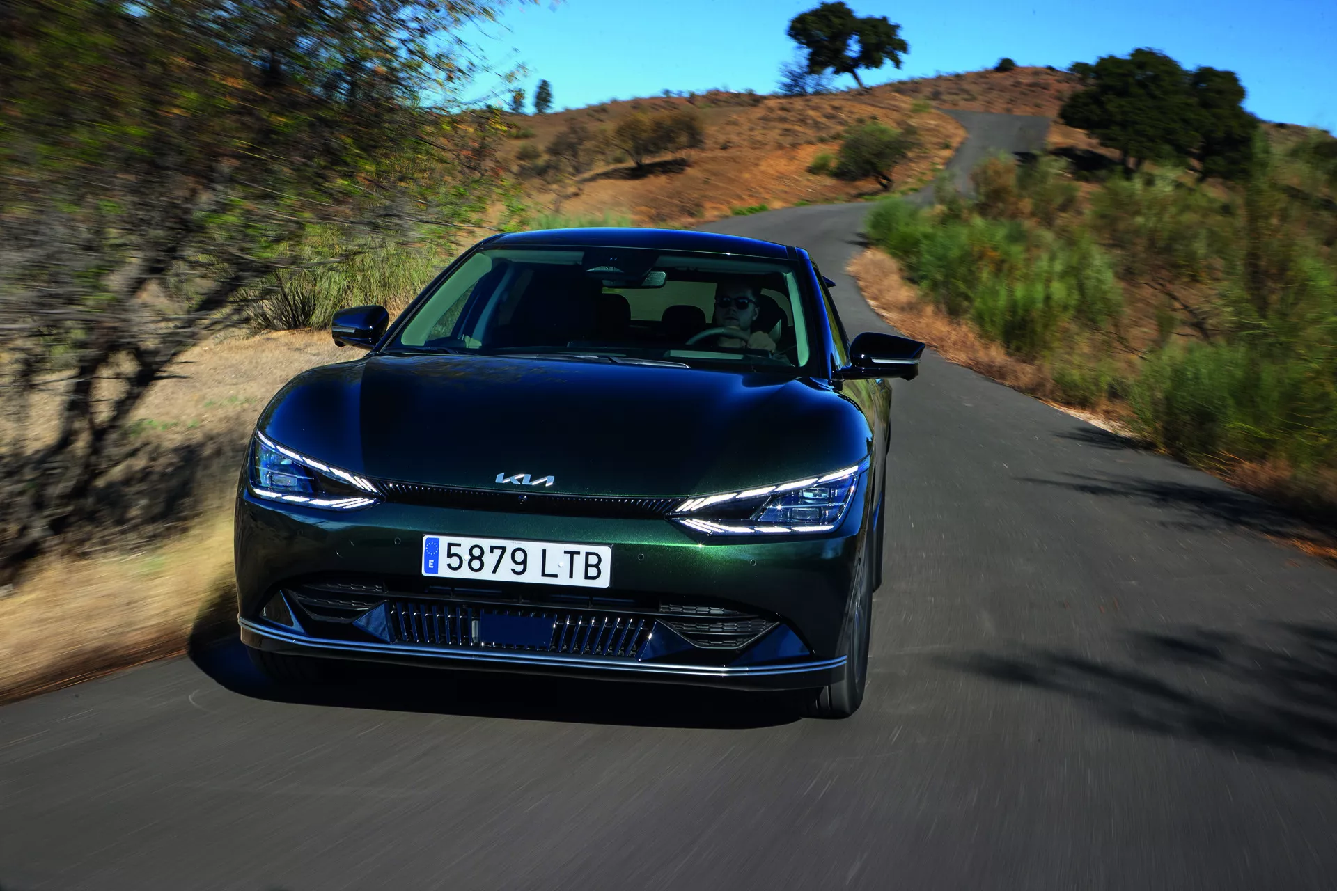 Dynamický pohyb Kia EV6 z roku 2021, pohľad z predného uhla zdôrazňujúci štíhle LED svetlomety, uzavretý dizajn mriežky a pokročilú technológiu asistencie vodiča na prírodnej scénickej ceste