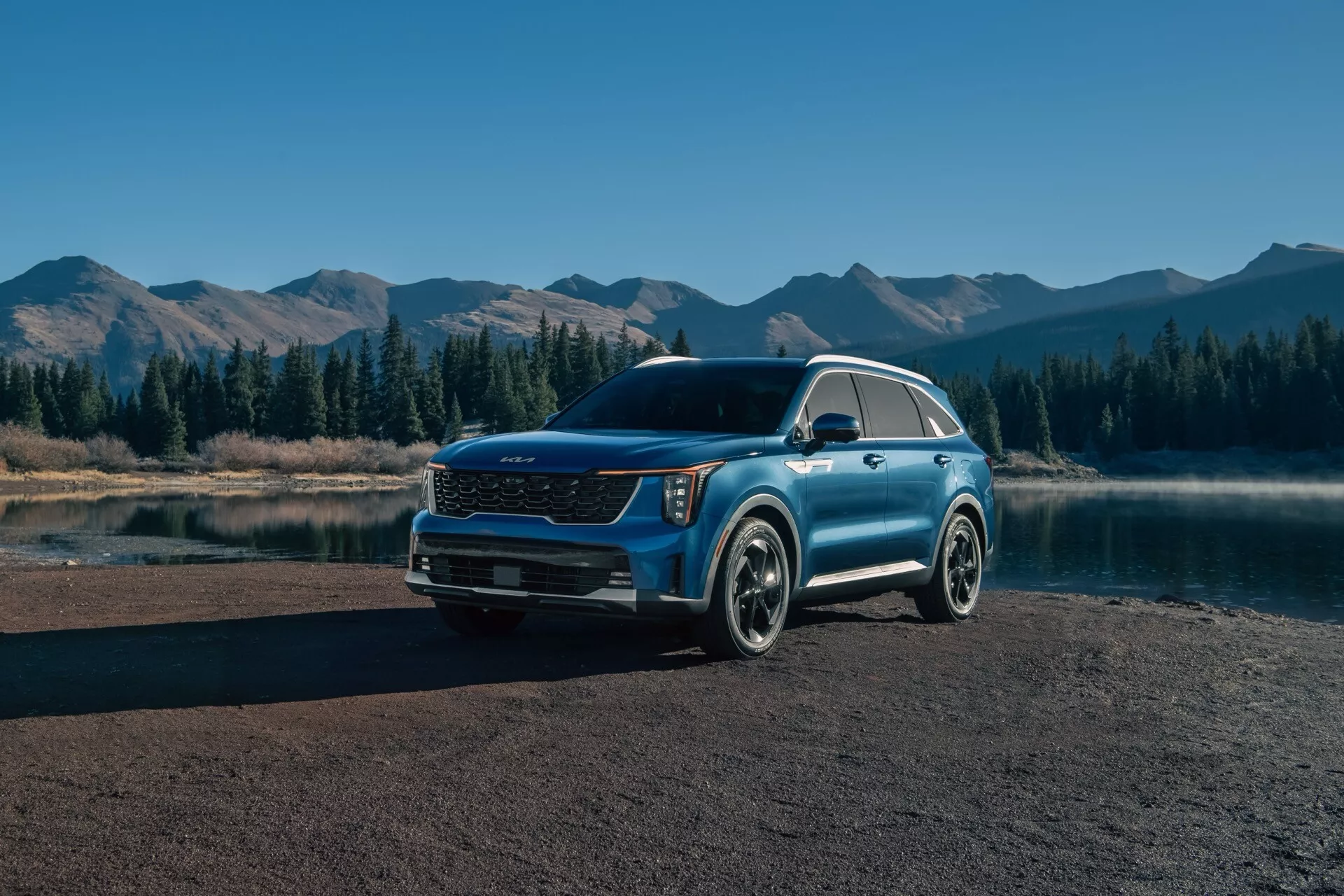 Kia Sorento MQ4-Facelift 2024 zaparkované v malebnom vonkom prostredí, zobrazujúce z trojštvrťového predného pohľadu, ktorý zdôrazňuje modernú prednú mriežku, LED svetlomety, metalízový lak a zliatinové kolesá na pozadí pokojného horského scenérie s odrazmi vody