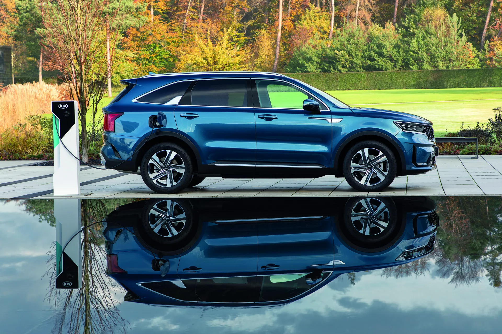 Moderné modré SUV Kia Sorento MQ4 2020 zaparkované na sivej dlažbe vedľa stanice pre nabíjanie elektrických vozidiel, zobrazuje bočný profil s čiastočným pohľadom spredu, zvýraznené prirodzeným ranným alebo večerným svetlom s pozadím zelenej listovej, zdôrazňuje schopnosť elektrického nabíjania, aerodynamický dizajn a disky z ľahkých zliatin.