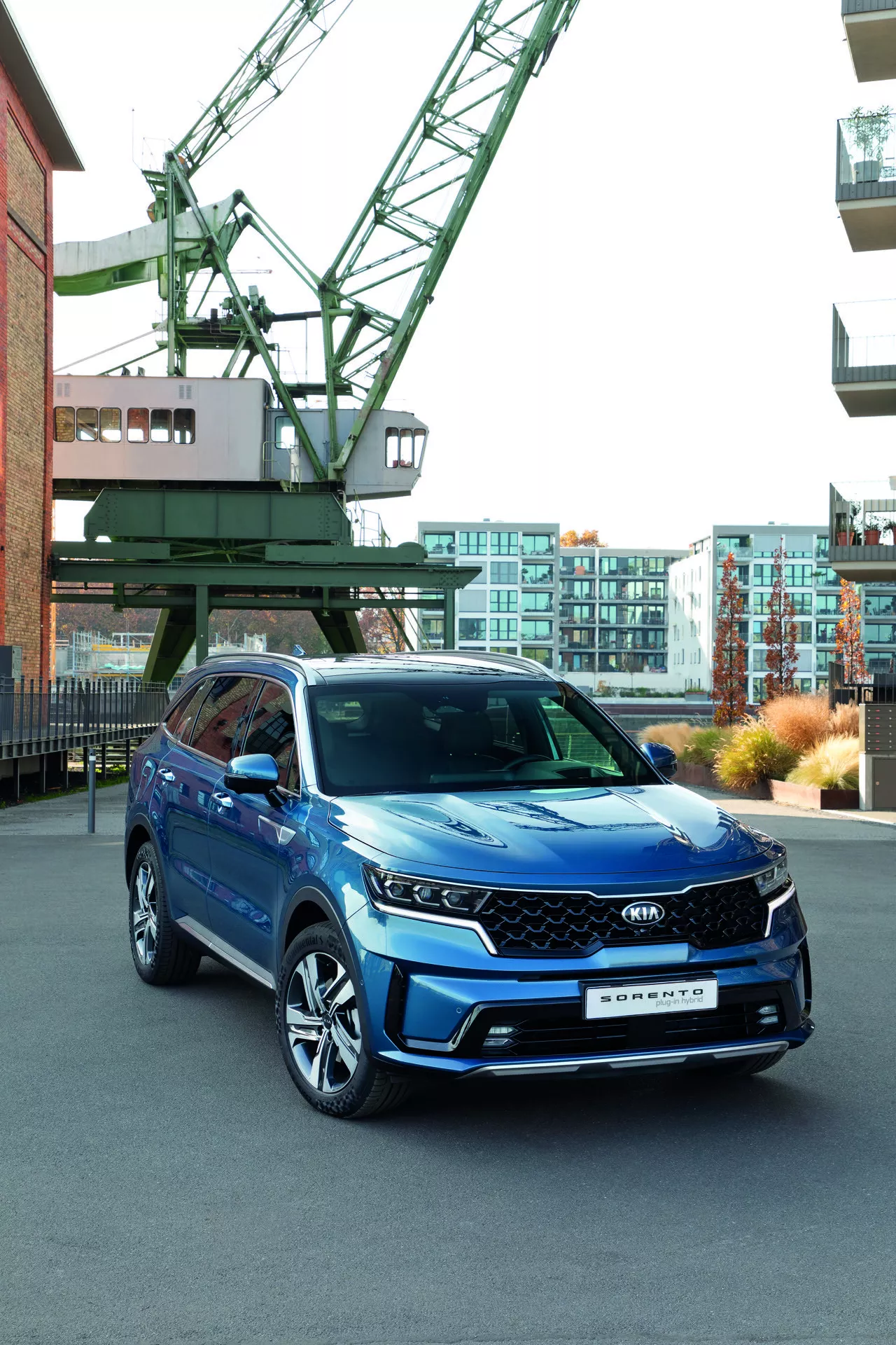 Modré Kia Sorento MQ4 z roku 2020 predstavené v mestskom prostredí s priemyselným žeriavom a modernými budovami na pozadí, s dôrazom na LED svetlomety, geometrickú mriežku s možnými senzormi ADAS, viacramenné zliatinové kolesá a integrované smerové signály v bočných zrkadlách, zdôrazňujúce jeho technologické vlastnosti a športový vzhľad.