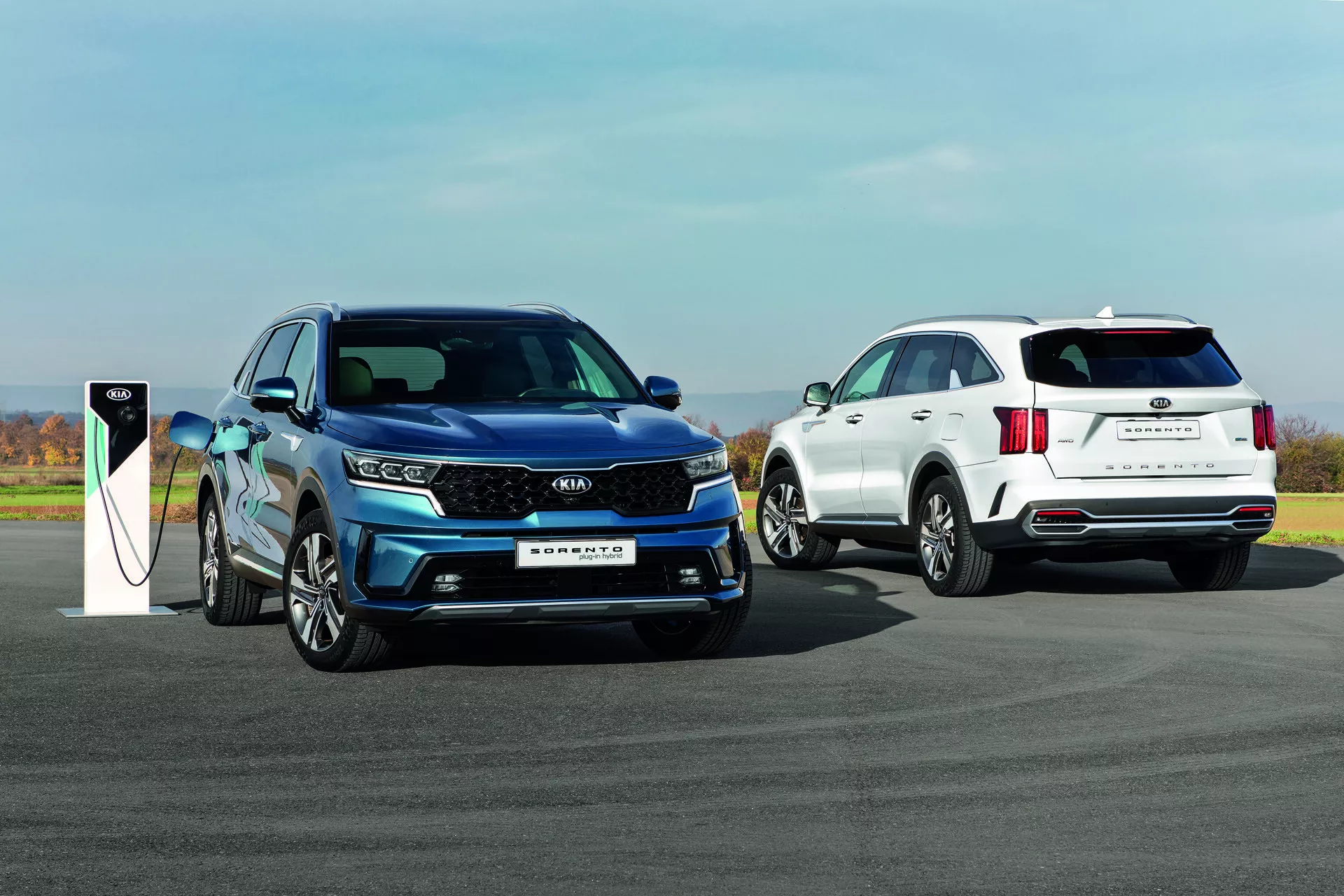 Kia Sorento MQ4 2020 v modrej farbe, model na elektrický pohon alebo plug-in hybridný model nabíjaný, a biely SUV na pozadí na spevnenej vonkajšej ploche pod jasnou oblohou