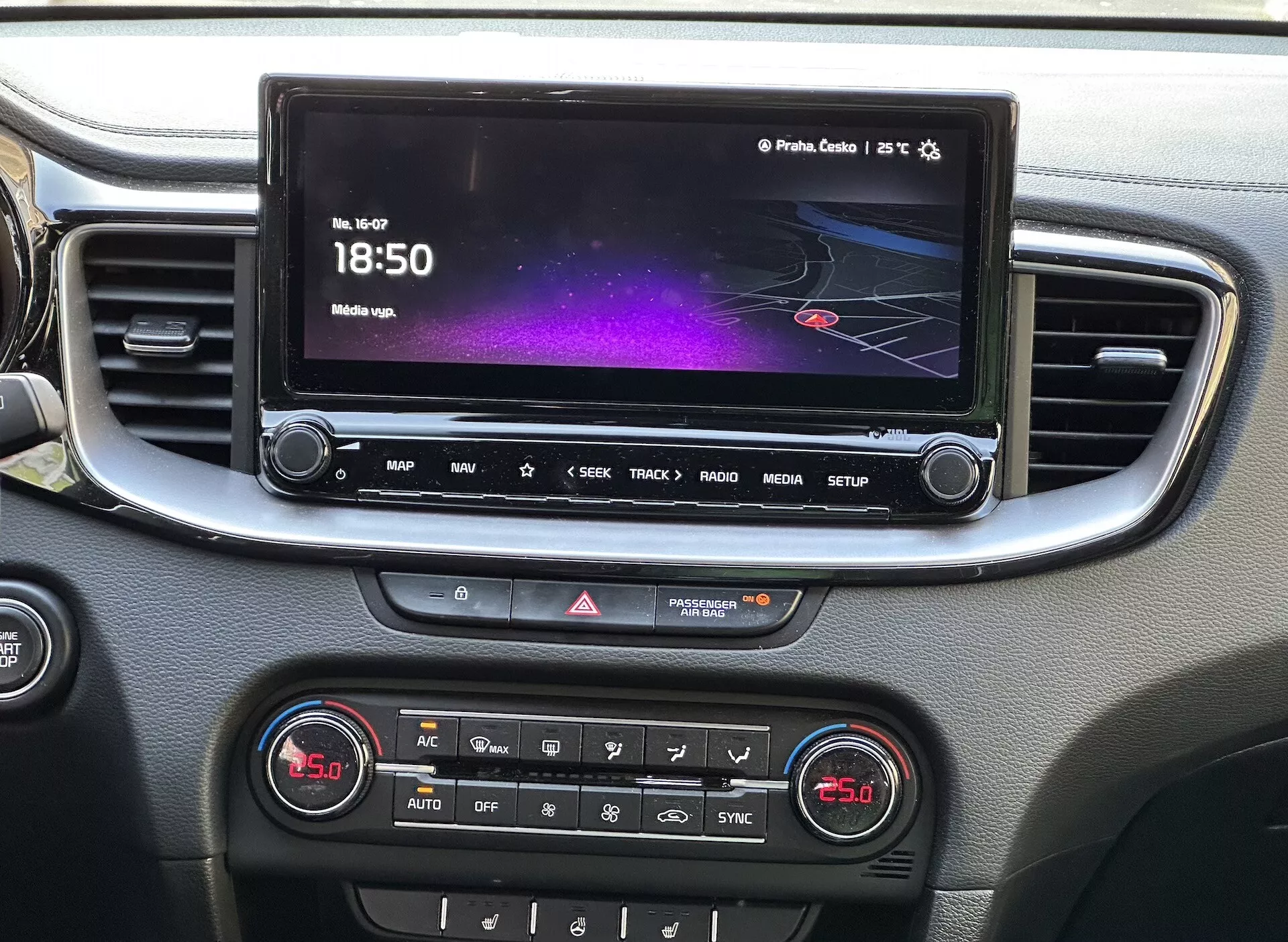 Podrobný pohľad na interiér Kia Xceed 2022 zobrazujúci stredovú konzolu s dotykovou obrazovkou infotainmentu, ktorá zobrazuje farebné rozhranie s navigáciou a informáciami o médiách, rýchlý prístup prostredníctvom radu tlačidiel, ovládanie varovných svetiel a panel s dvojzónovou klimatizáciou, všetko v ráme so striebornou líniou.