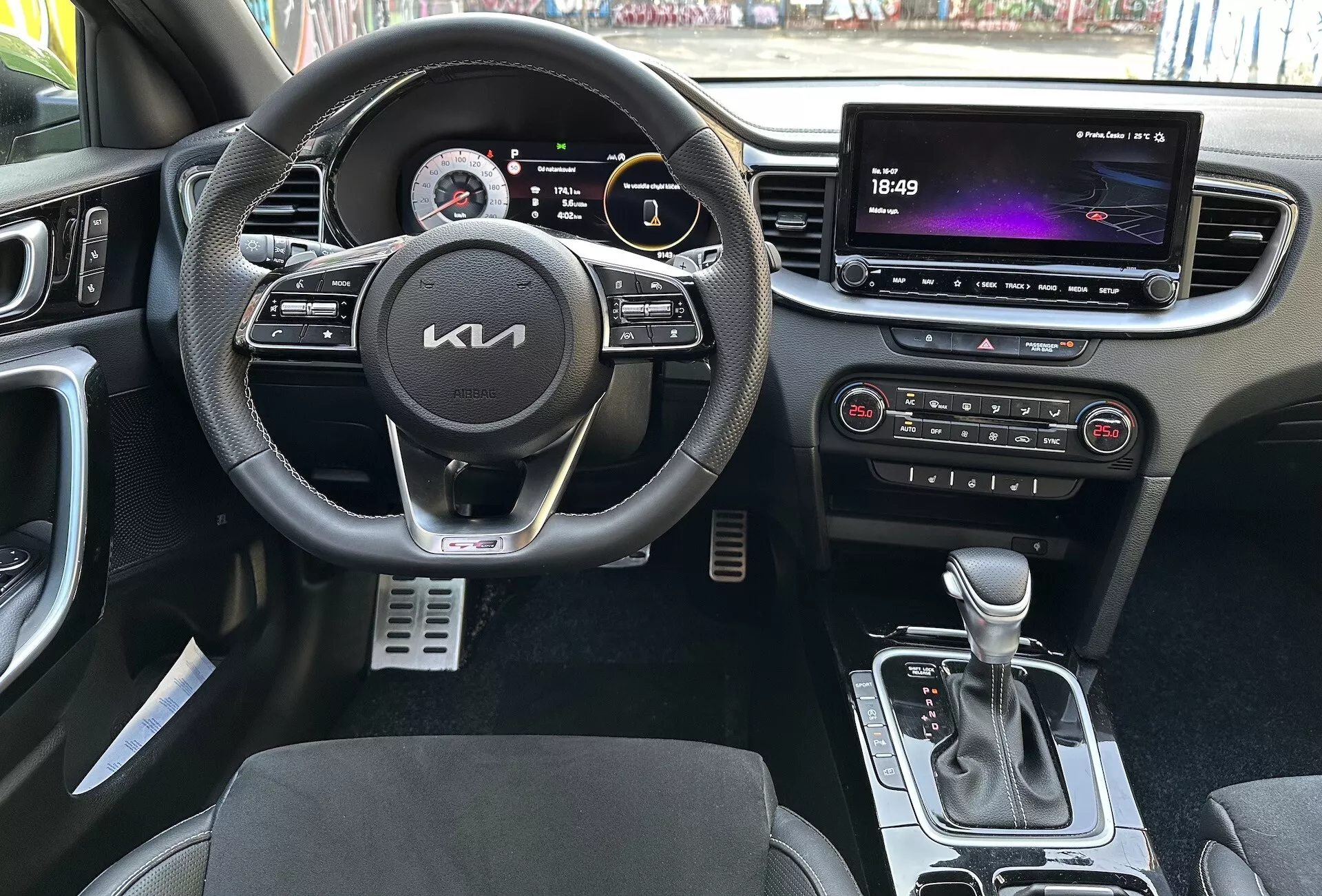 Interiérový pohľad na Kia XCeed 2022 zdôrazňujúci funkcie zamerané na vodiča, ako je multifunkčný volant obalený kožou s tlačidlami pre multimédiá a tempomat, digitálny prístrojový panel, veľký dotykový informačno-zábavný systém, automatické ovládanie klímy, automatická radiaca páka, sedadlá s dvojfarebným čalúnením a kovové športové pedále z perspektívy prvej osoby.