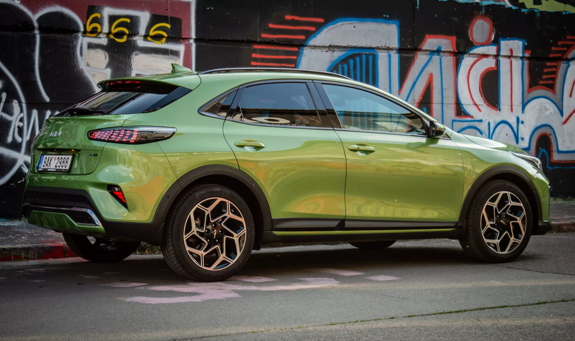 Pohľad z boku na Kia XCeed 2022 zdôrazňujúci dizajn zadného kolesa, metalickú zelenú farbu, LED zadné svetlá a štýl kompaktného SUV s aerodynamickými prvkami na pozadí mestského street artu.