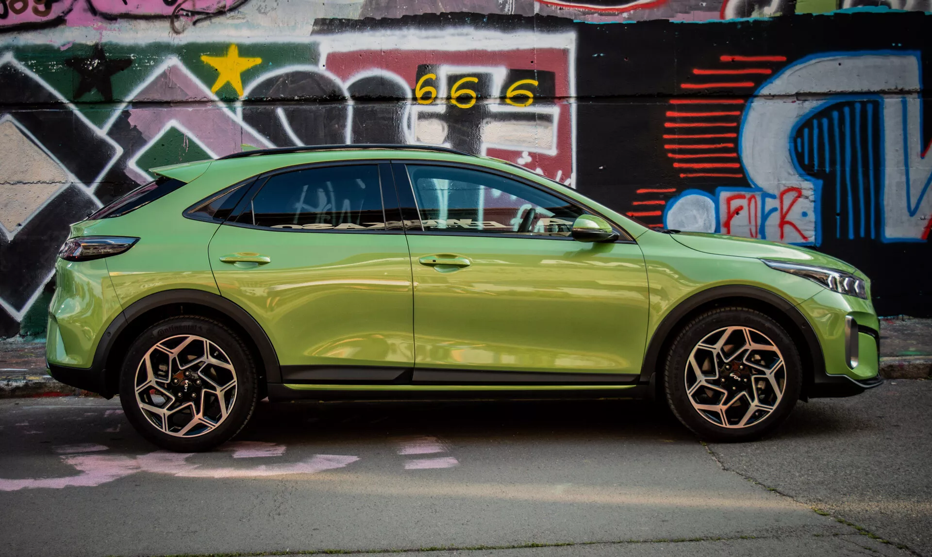 Kompaktné SUV Kia XCeed 2022 v limetkovej zelenej farbe zaparkované pri stene pokrytej graffiti, predvádzajúce svoj elegantný dizajn a pokročilé funkcie ako je bezkľúčový prístup a integrované smerové signály v bočných zrkadlách.