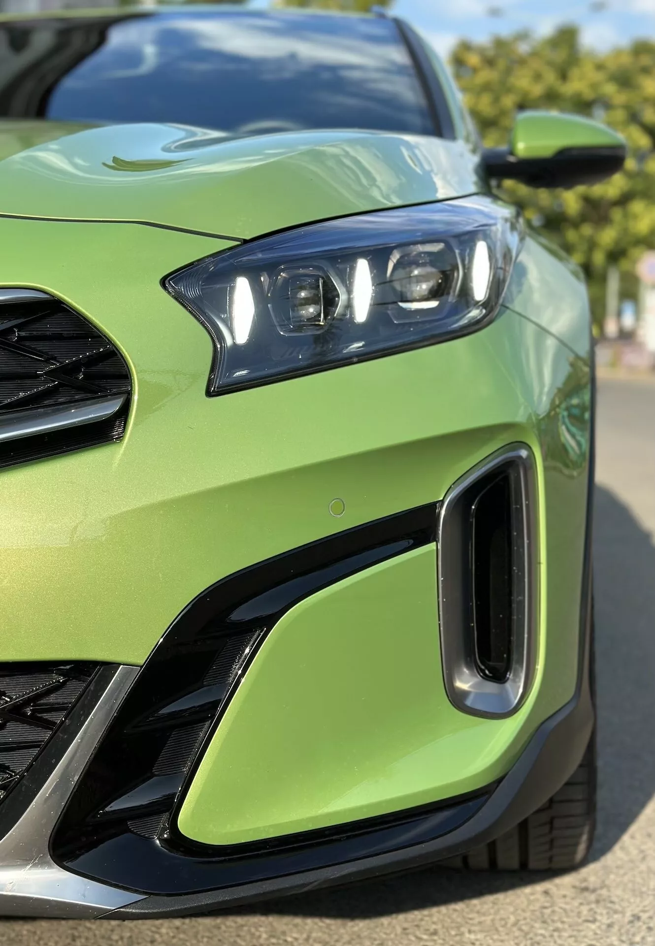 Detailný záber na prednú ľavú štvrtinu Kia Xceed 2022 ukazujúci súpravu LED svetlometov, časť mriežky, vzduchové vstupy na prednom nárazníku, proti limetkovo zelenej farbe karosérie, zdôrazňujúci športový dizajn vozidla a pokročilú svetelnú technológiu.