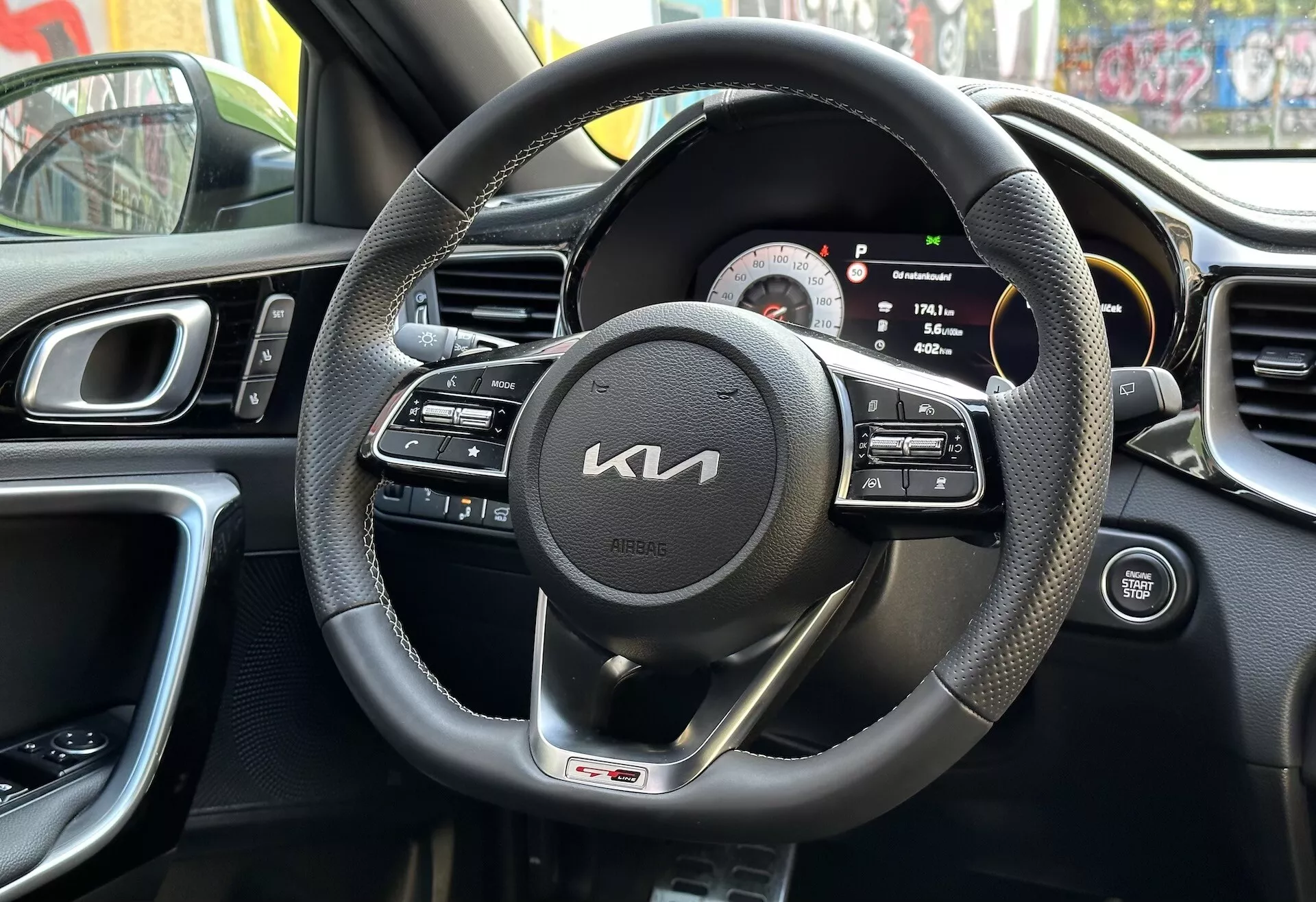Interiér Kia XCeed 2022 z pohľadu vodiča s čiernym volantom obaleným kožou s multifunkčnými ovládačmi a odznakom GT línie, digitálno-analógovým prístrojovým panelom, časťou stredovej konzoly s ventiláciami, ergonomickým dizajnom palubnej dosky a panelom dverí vodiča s ovládaním, odrážajúci mestské prostredie