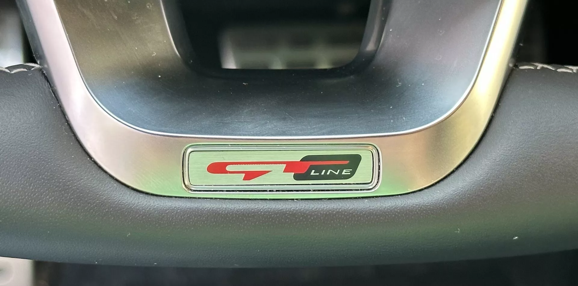 Detailný záber na emblém volantu Kia XCeed 2022 GT-Line s kovovými a čiernymi písmenami a športovým červeným akcentom, obklopeným prémiovým volantom z kože a kovu