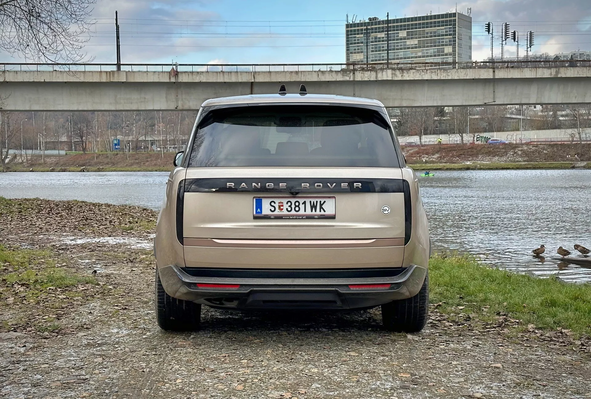 Zadný pohľad na Range Rover 2021 s priamym uhľom pohľadu na úrovni očí, zaparkovaný blízko vodného tela s kačkami a nadchodom na pozadí, zobrazujúci názov modelu, zadné svetlá, parkovacie senzory a dvojitý výfukový systém v matnej béžovej farbe
