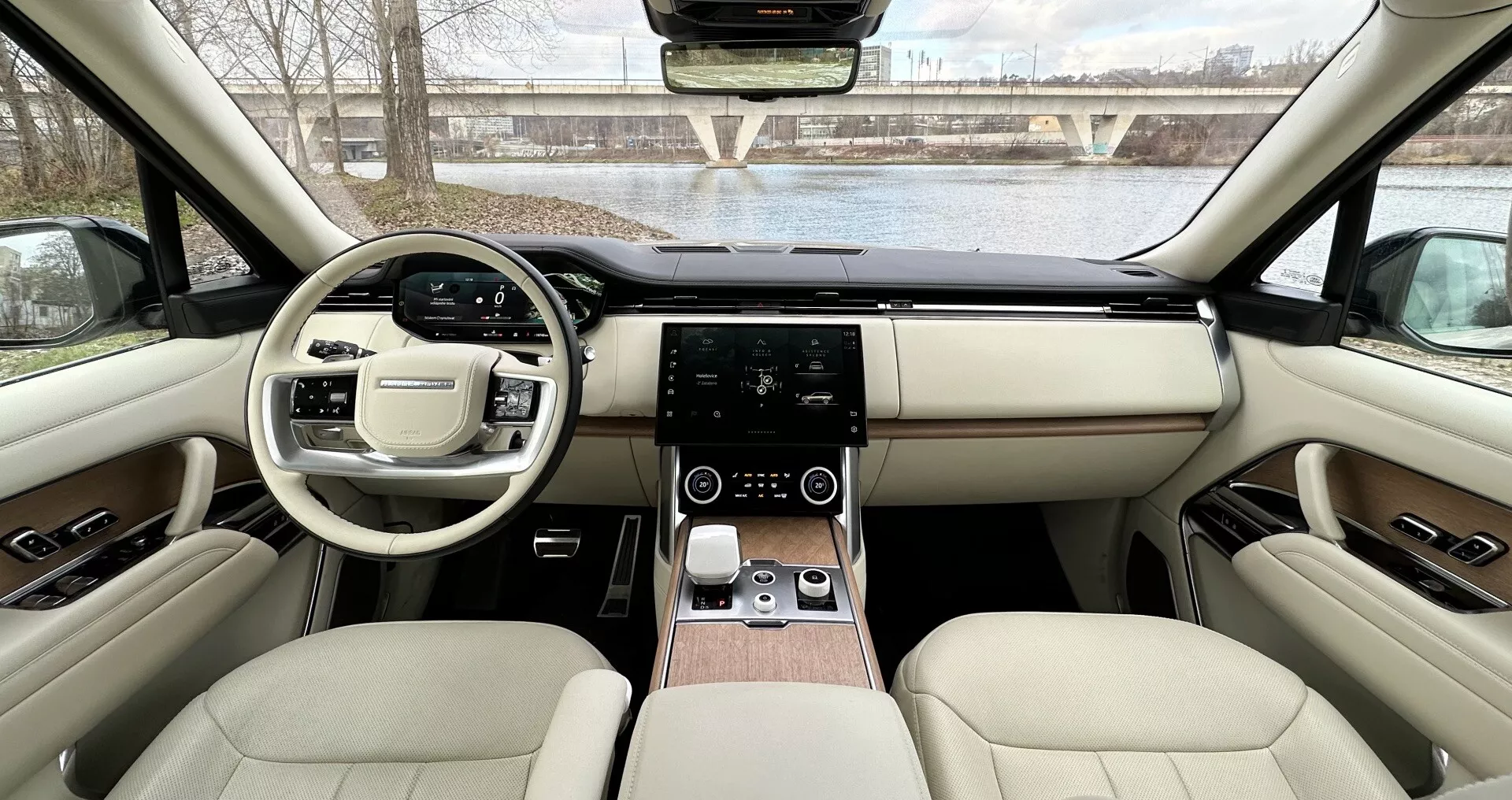 Interiér Land Rover Range Rover 2021 z pohľadu zo zadných sedadiel smerom k prístrojovej doske ukazujúci symetrický dizajn, volant s multifunkčnými tlačidlami, dotykový infotainment systém, digitálne ovládanie klímy, luxusný kožený poťah s drevitými akcentmi a pokročilé technologické funkcie