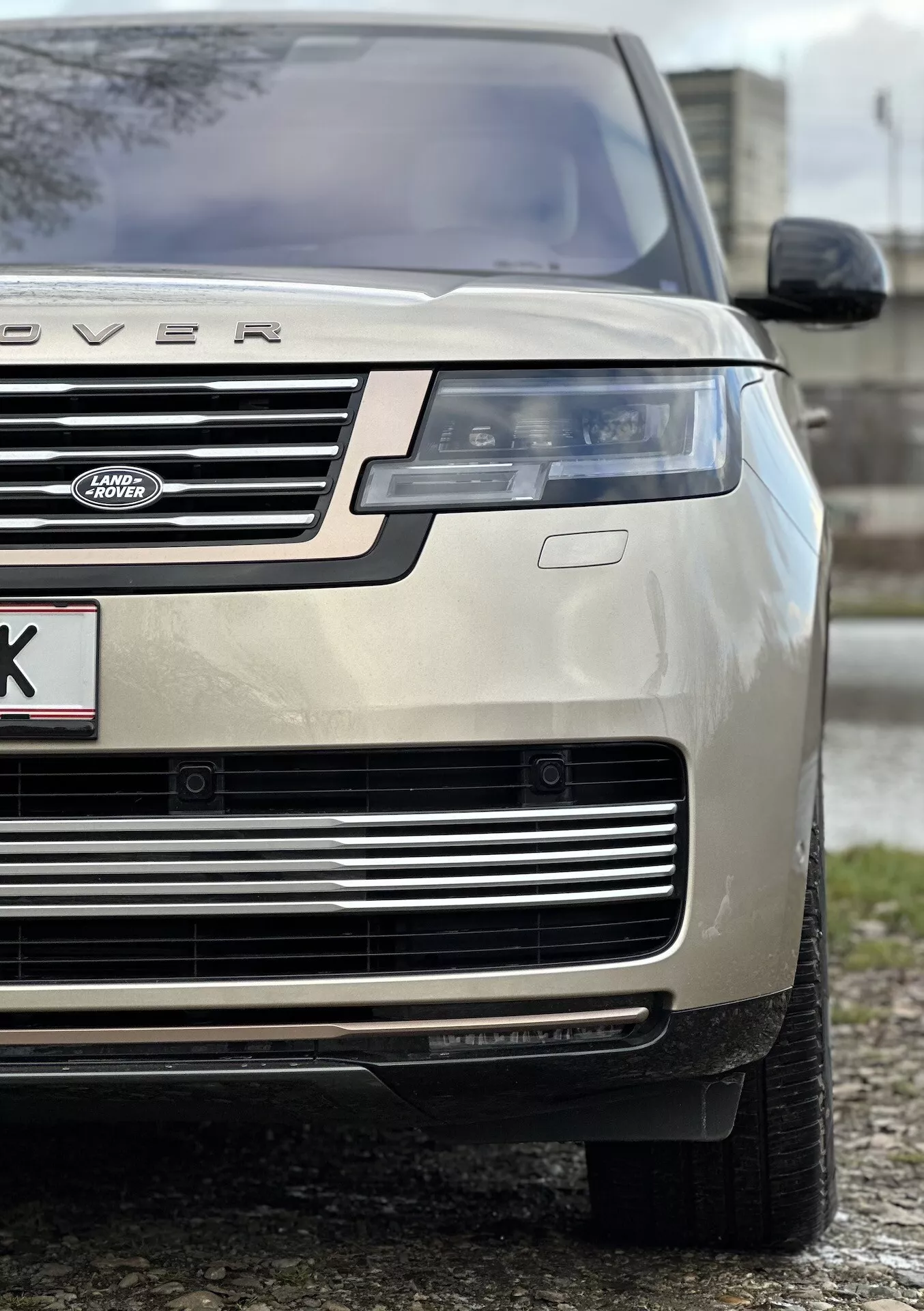 Detailný záber zlatého Land Rover Range Rover 2021 na prednú ľavú časť vozidla, zdôrazňujúci výšku a postoj vozidla so zameraním na kapotu, mriežku, LED svetlomet, nárazník a celoročnú pneumatiku, naznačujúci luxus a schopnosť terénnej jazdy.