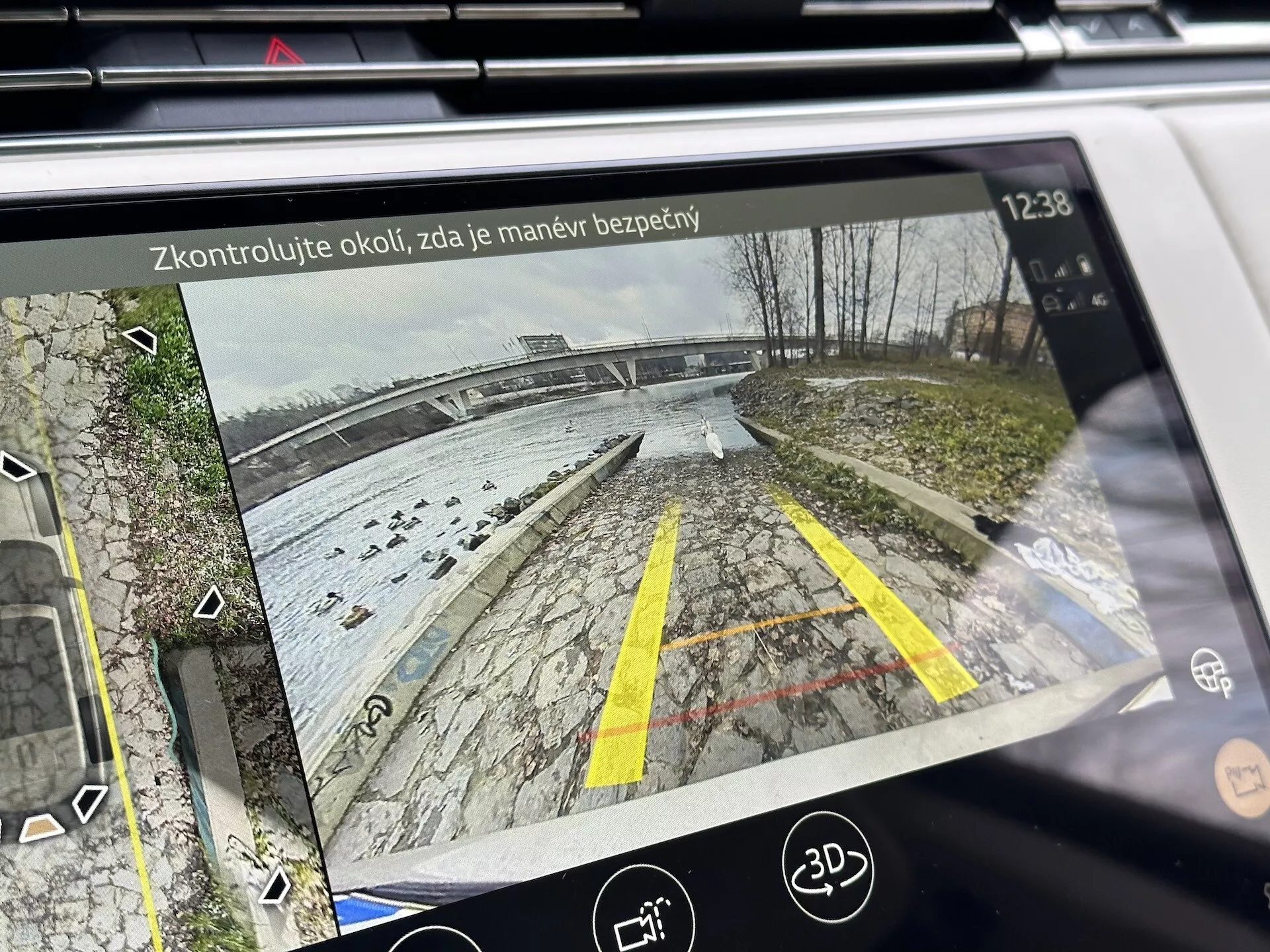 Displej infotainment systému Land Rover Range Rover 2021 zobrazujúci zadný pohľad z kamery pre cúvanie s dynamickými vodiacimi čiarami na parkovisku pri vodnej ploche, detaily s vegetáciou, ikonami asistencie pri parkovaní a bezpečnostným upozornením