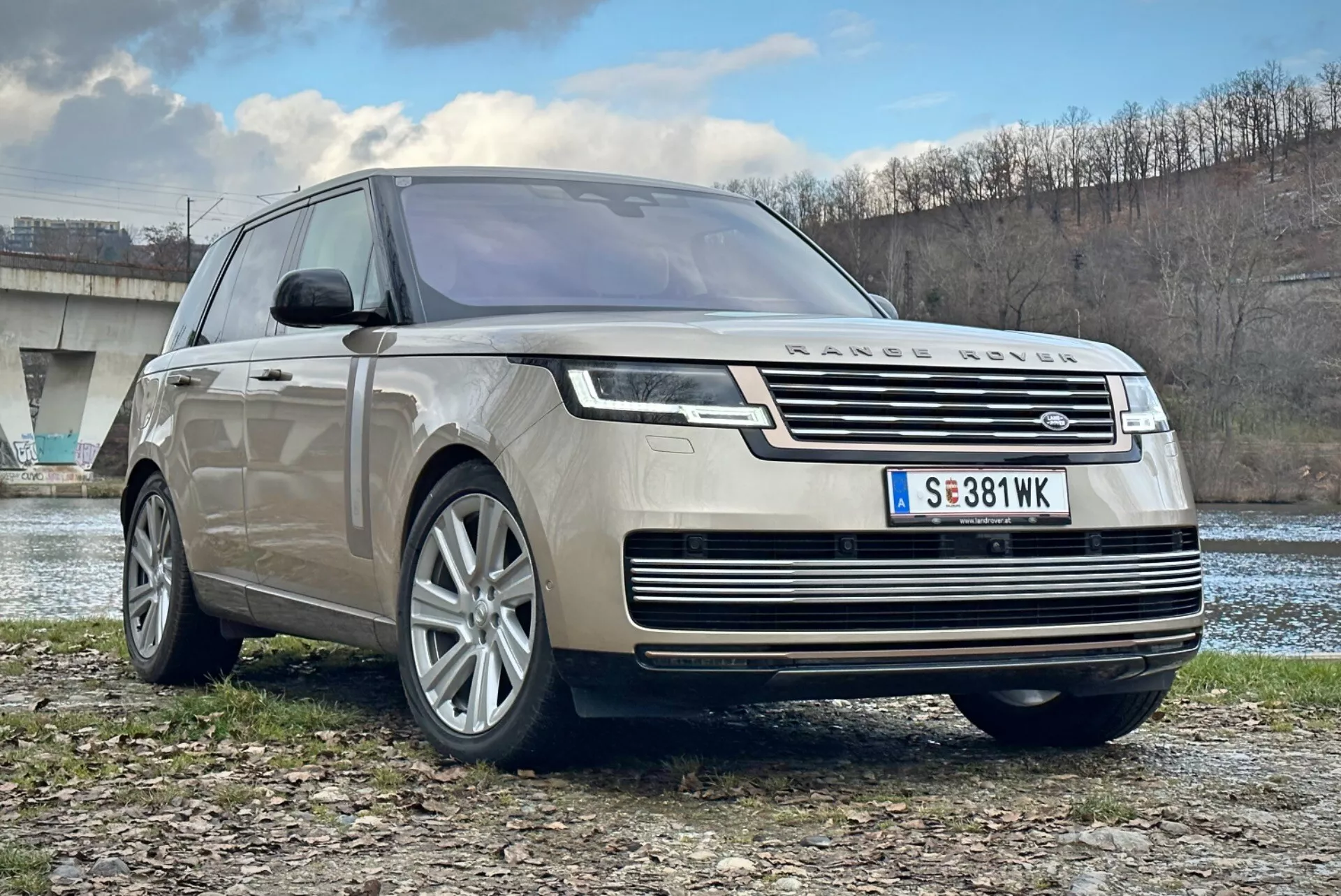 Luxusné SUV Land Rover Range Rover 2021, pohľad z prednej a trojštvrťovej strany zobrazujúci chrómovanú mriežku, LED svetlomety, viacdielne zliatinové kolesá a aerodynamický dizajn, zaparkované vonku blízko rieky a mostu
