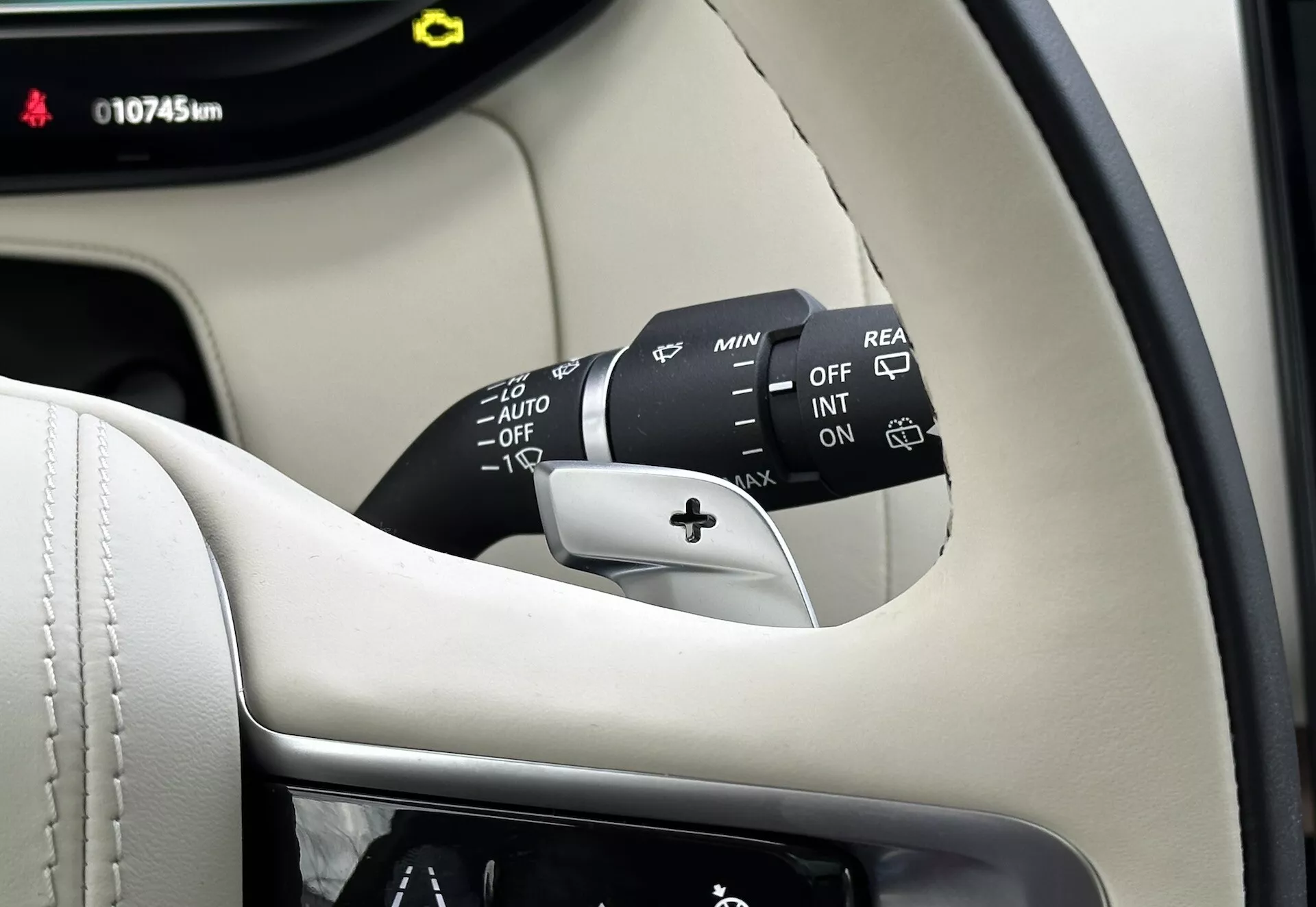 Detailný pohľad dovnútra Land-Rover Range-Rover 2021 zobrazujúci stĺpik riadenia, páčku riadenia stieračov s viacerými nastaveniami vrátane OFF, INT, ON, regulátory rýchlosti a AUTO pre automatické stierače s dažďovým senzorom, plus možnosť zadného stierača. Viditeľná časť prístrojového panela zobrazuje 10745 km, všetko proti pozadiu prémiového čierneho a smotanového interiéru.