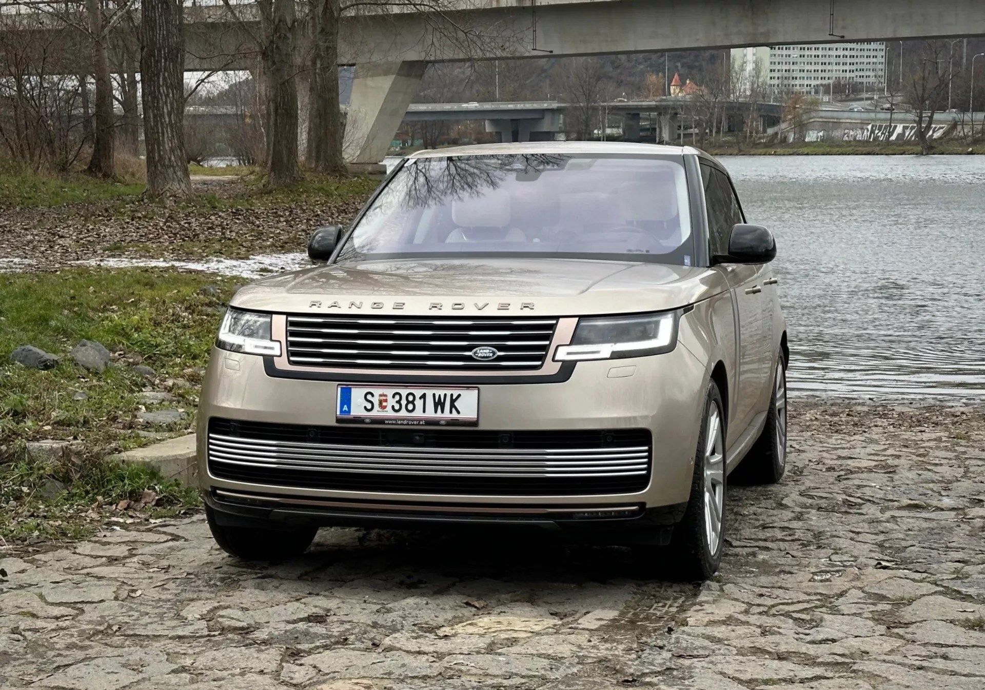 Luxusné SUV Range Rover 2021 v striebornej farbe, zvýrazňuje prednú mriežku s brandovaním, štíhlé LED denné svetlá, predný nárazník s parkovacími senzormi a dizajn kapoty clamshell na dlažobnej ploche blízko rieky