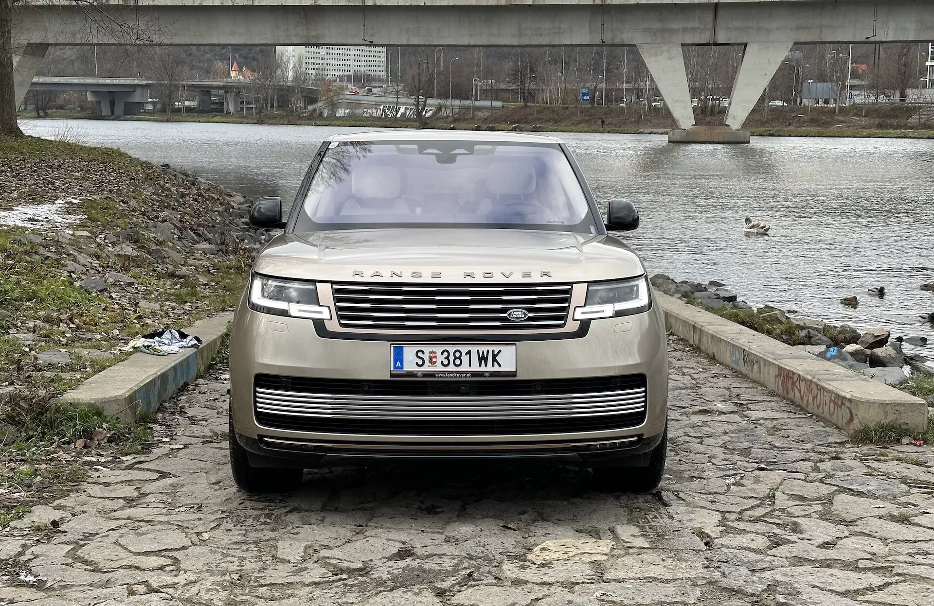 Land Rover Range Rover 2021 zobrazený z prednej, mierne nízkej uhla zvýrazňujúceho mriežku, predný nárazník a kapotu, s ikonickým emblémom RANGE ROVER, modernými LED svetlometmi a pokročilou technológiou asistencie vodiča, na pozadí rieky alebo jazera, mosta a zamračenej oblohy