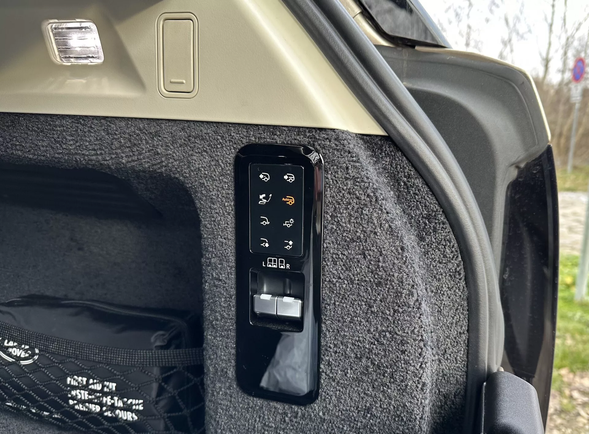 Detailný pohľad na elektronický ovládací panel kufra vo vnútri Land Rover Range Rover 2021, zobrazujúci tlačidlá pre operácie ako elektronické otváranie a zatváranie, ovládanie skladacieho nákladového krytu, potenciálne svetlo kufra alebo pomocnú zásuvku, nastavenie polohy zadných sedadiel z kufra a núdzové uvoľnenie, umiestnené proti kobercovému bočnému stenu kufra a svetlému stropu s nákladovým svietidlom.