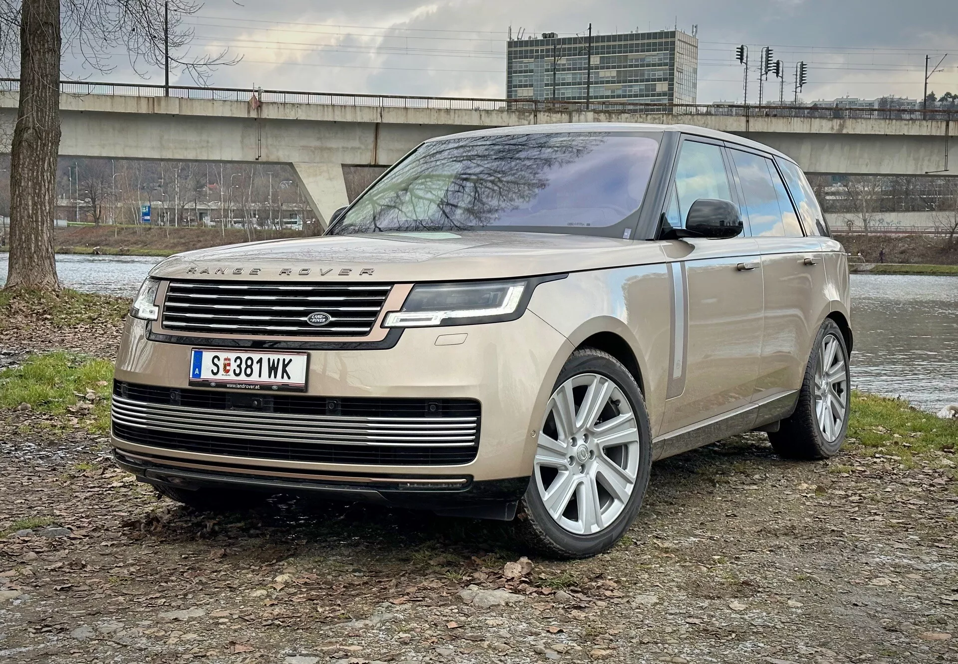 Luxusné SUV Land Rover Range Rover 2021 zaparkované na zmiešanom teréne zeme a trávy s betónovou štruktúrou a vegetáciou na pozadí, zobrazujúce pohľad z predného uhla s dôrazom na LED svetlomety, zliatinové kolesá a moderné aerodynamické prvky