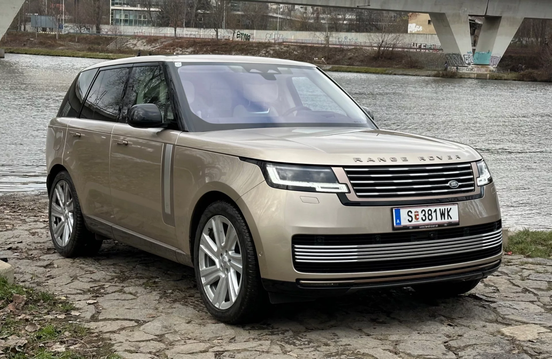 Luxusné SUV Land Rover Range Rover ročníku 2021 zaparkované na brehu rieky s mostom na pozadí, ukazujúce jeho moderný dizajn a technologické prvky vrátane LED svetlometov, prednej mriežky s emblémou, viacramenných zliatinových kolies a panoramatického strešného okna.