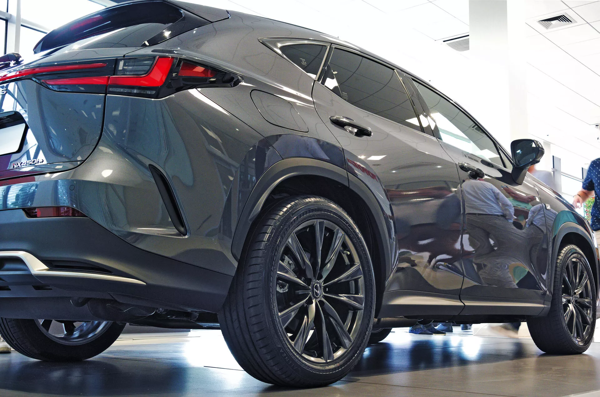 Zadný trojštvrťový uhol vozidla Lexus NX 2021 SUV ukazujúci bočný profil a zadnú časť s veľkými viacramennými zliatinovými kolesami, nízkoprofilovými pneumatikami, ostrými zadnými LED svetlami, kovovým lakom, tónovanými oknami, integrovaným zadným spojlerom a vylešteným showroomovým prostredím zdôrazňujúcim modernú estetiku a luxusné prvky
