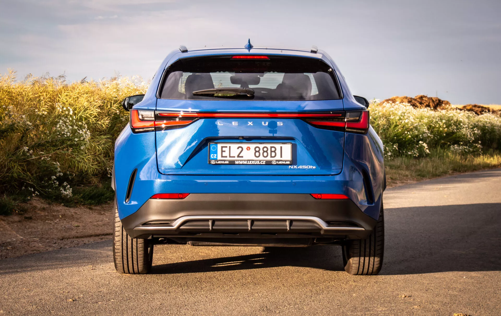 Zadný pohľad na modrý Lexus NX 2021 z nízkeho uhla zvýrazňujúci pevnú stavbu a dizajnové prvky auta ako sú LED zadné svetlá, zadné čelné sklo, spoiler, výrazné značenie, zadný nárazník s difúzorom, parkovacie senzory, odrazky a dvojitý výfuk na pozadí prírody pod jasným nebom.