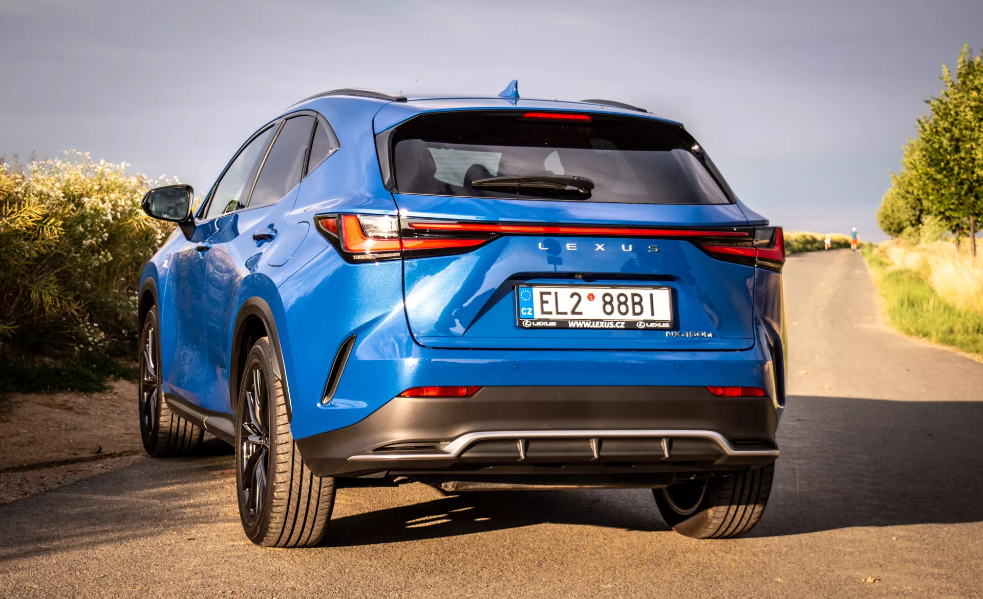 Zadný pohľad na luxusné crossover SUV Lexus NX modrej farby ročník 2021, ktoré predvádza LED zadné svetlá, značenie Lexus, športový zadný nárazník s difúzorom, dvojité výfukové rúry a ďalšie technologické a dizajnové prvky z nízkeho uhla a trojštvrtečnej perspektívy