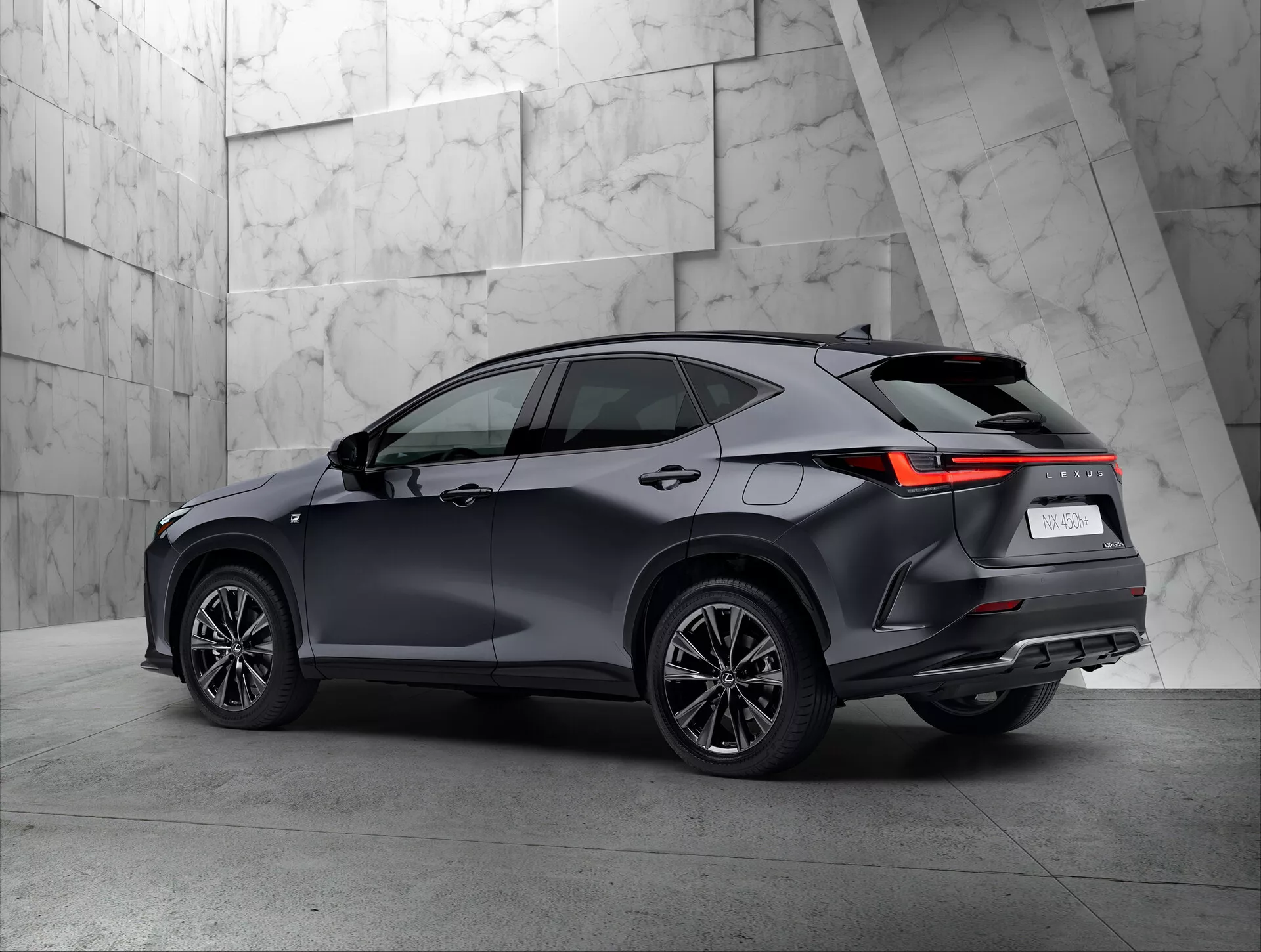 Trojštvrťový zadný pohľad na moderné SUV Lexus NX 2021 v showroome. Vozidlo zreteľne zobrazuje pravú stranu a zadnú časť, vrátane zadných svetiel s LED dizajnom, tmavým lesklým lakom, viacramennými kolesami a strechou s možnými strešnými nosičmi. Neutrálne, textúrované pozadie pekne kontrastuje so sofistikovaným dizajnom SUV.