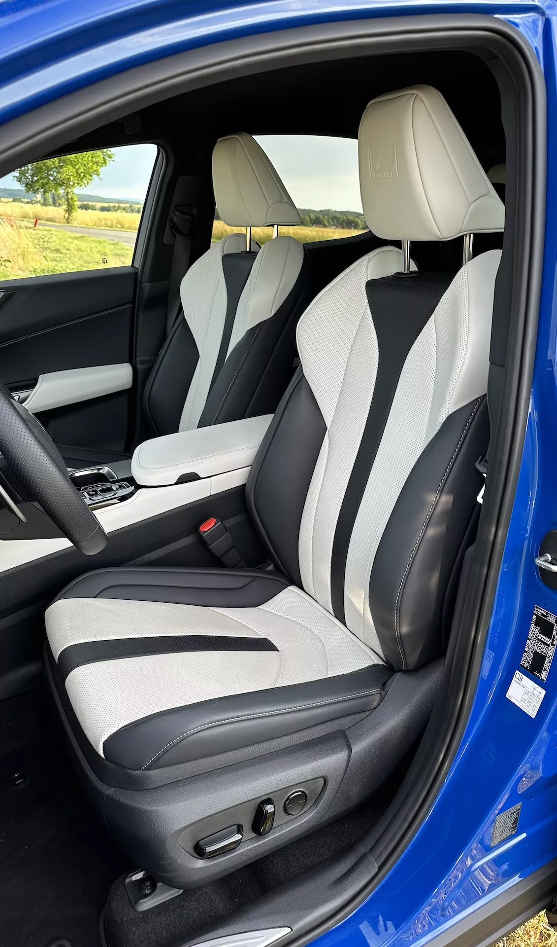 Pohľad do interiéru cez otvorené dvere vodiča Lexus NX 2021, zobrazujúci oblasť vodiča z miernych výšok, s dvojfarebným koženým čalúnením, pokročilými ovládacími prvkami sedadla vodiča, bočným spevnením, integrovanými opierkami hlavy a centrálnou konzolou s automatickou pákou prevodovky