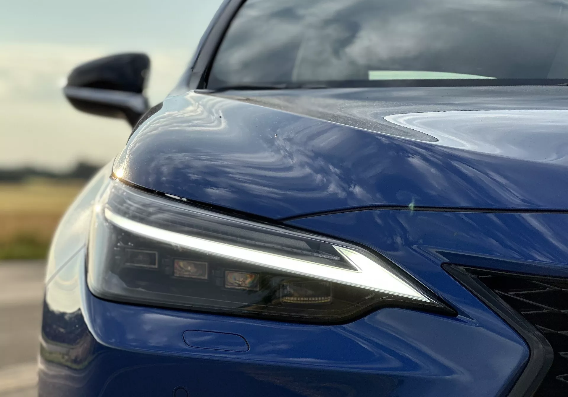 Detailný pohľad na ľavú prednú štvrtinu Lexusu NX 2021 s LED svetlometom, tvarovanou kapotou, predným nárazníkom s mriežkou a lesklým tmavo modrým povrchom, zdôrazňujúci pokročilú technológiu osvetlenia a aerodynamický dizajn.