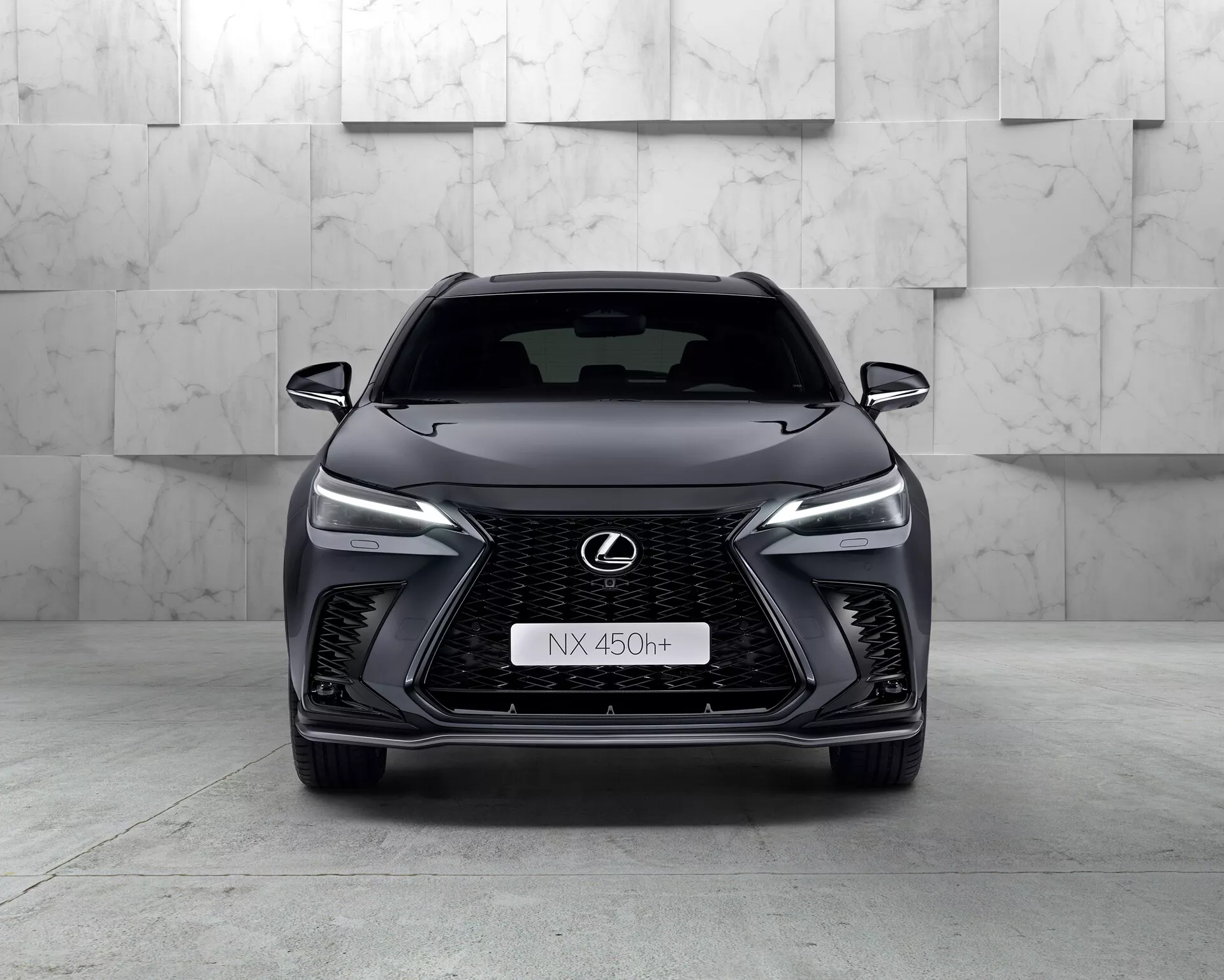 Čelný pohľad na Lexus NX 450h+ z roku 2021, elegantné moderné SUV, ktoré ukazuje svoju výraznú vretenovitú masku s mriežkovým vzorom, zúžené LED svetlomety, logo Lexus s modrým obrysom a agresívny dizajn predného nárazníka. Hybridný charakter vozidla je indikovaný znakom 'h+' na maske. Štúdiové prostredie zvýrazňuje krivky a vlastnosti vozidla čistým, kontrolovaným osvetlením.