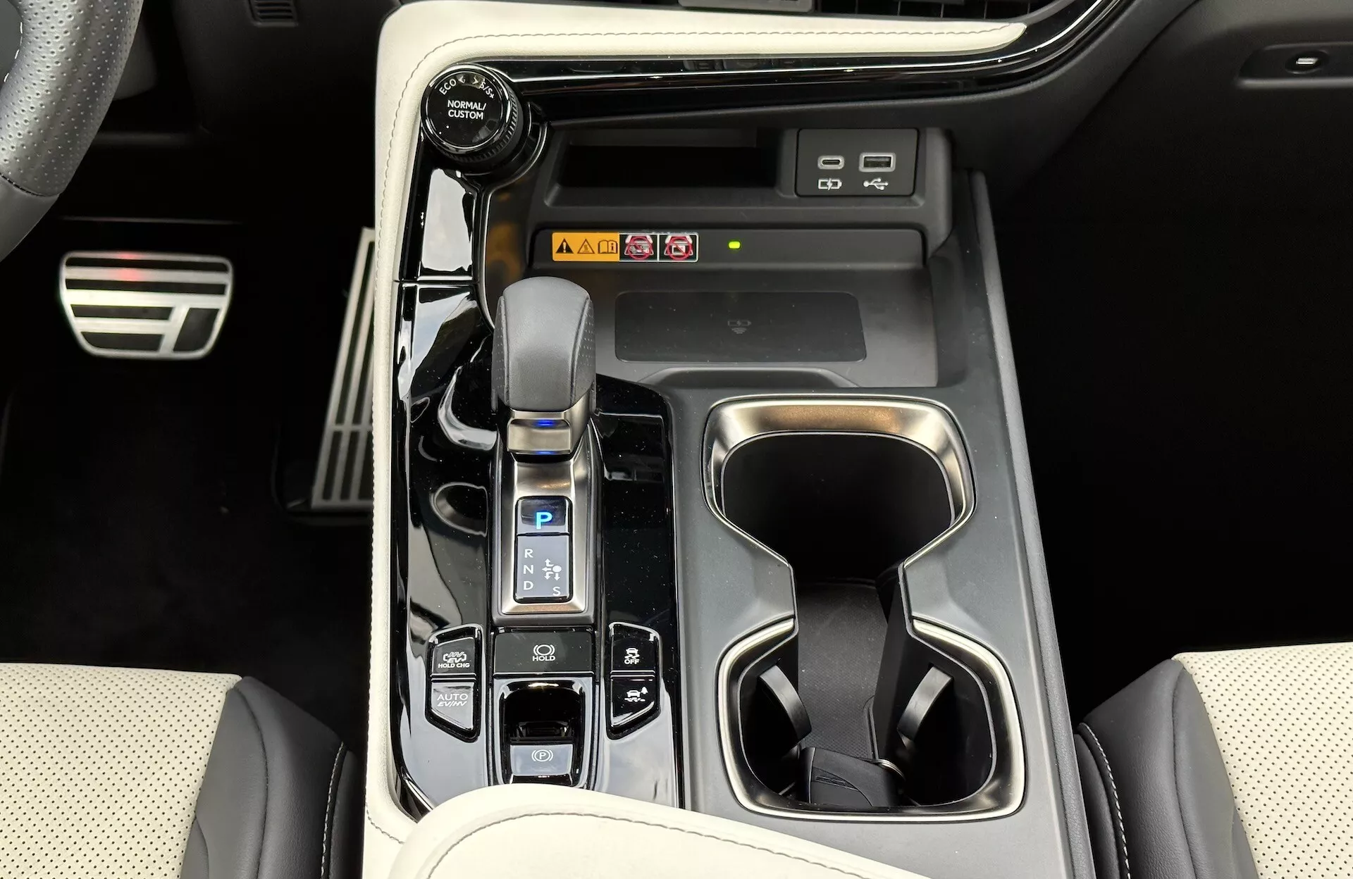 Pohľad na stredovú konzolu Lexus NX 2021 ukazujúci automatickú radiaciu páku s koženým manžetou, volič jazdných režimov, držiaky na nápoje, tlačidlá ovládania vozidla, potenciálne skratky pre informačno-zábavný systém a prvky luxusného dizajnu interiéru