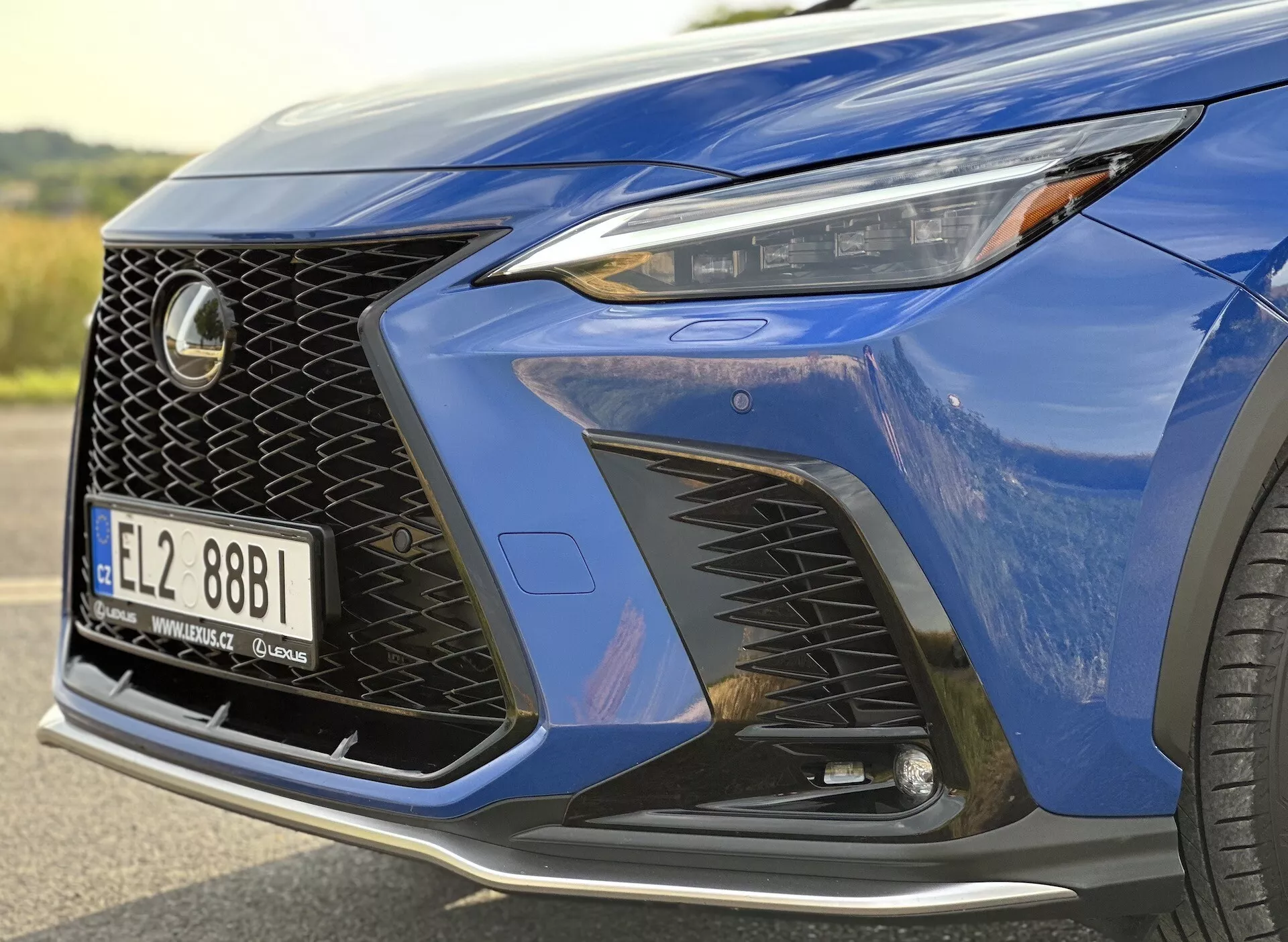 Detailný pohľad na predný roh Lexusu NX 2021 s predným svetlometom s technológiou LED, výrazným dizajnom mriežky chladiča a aerodynamickým predným nárazníkom s veľkými vstupmi vzduchu, zvýraznený metalízou hlboko modrej farby a náhľadom na luxusné zliatinové koleso.