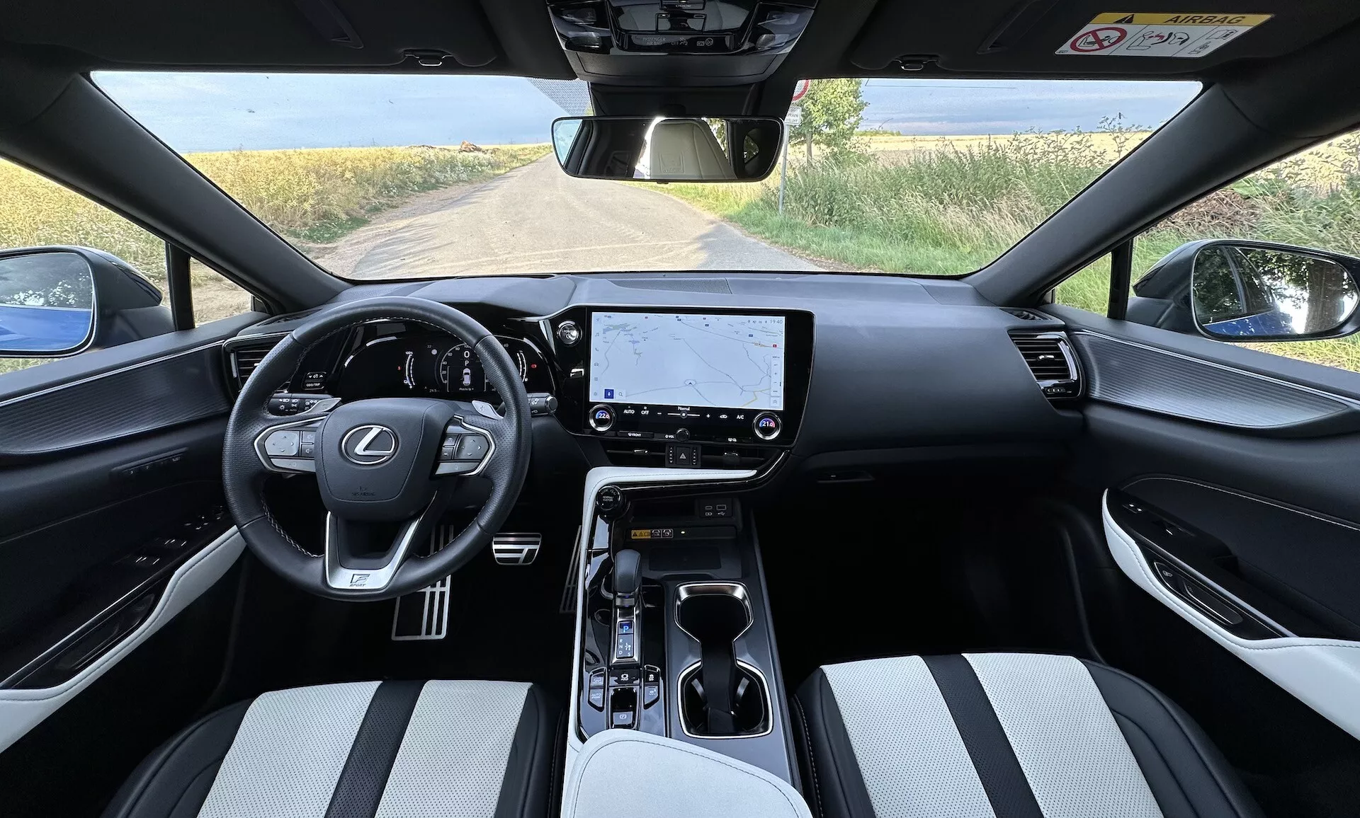 Interiér Lexusu NX 2021 z pohľadu vodiča s volantom uprostred s logom Lexus a ovládacími tlačidlami, veľkou dotykovou obrazovkou na palubnej doske, minimalistickými ovládačmi klímy, posuvnou automatickou prevodovkou na stredovej konzole s držiakmi na nápoje a úložným priestorom, sedadlá čalúnené dvojfarebným materiálom, a čiastočný výhľad na vidiecku krajinu cez čelné sklo.