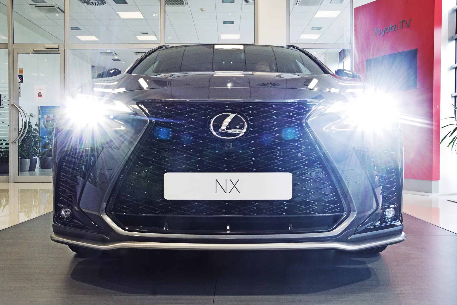Predný pohľad na vozidlo Lexus NX 2021 fotografované z nízkeho uhla, ktoré ukazuje jeho výšku a prítomnosť na ceste v prostredí showroomu. Obrázok zvýrazňuje charakteristickú vretenovú mriežku s tvarom presýpacích hodín a tmavou mriežkou, osvetlené uhlové LED svetlomety s efektmi iskrienia, denné svetlá a detaily spodného nárazníka. Lesklá podlaha a držiak poznávacej značky s nápisom 'NX' dodávajú vozidlu luxusný a technologický vzhľad.