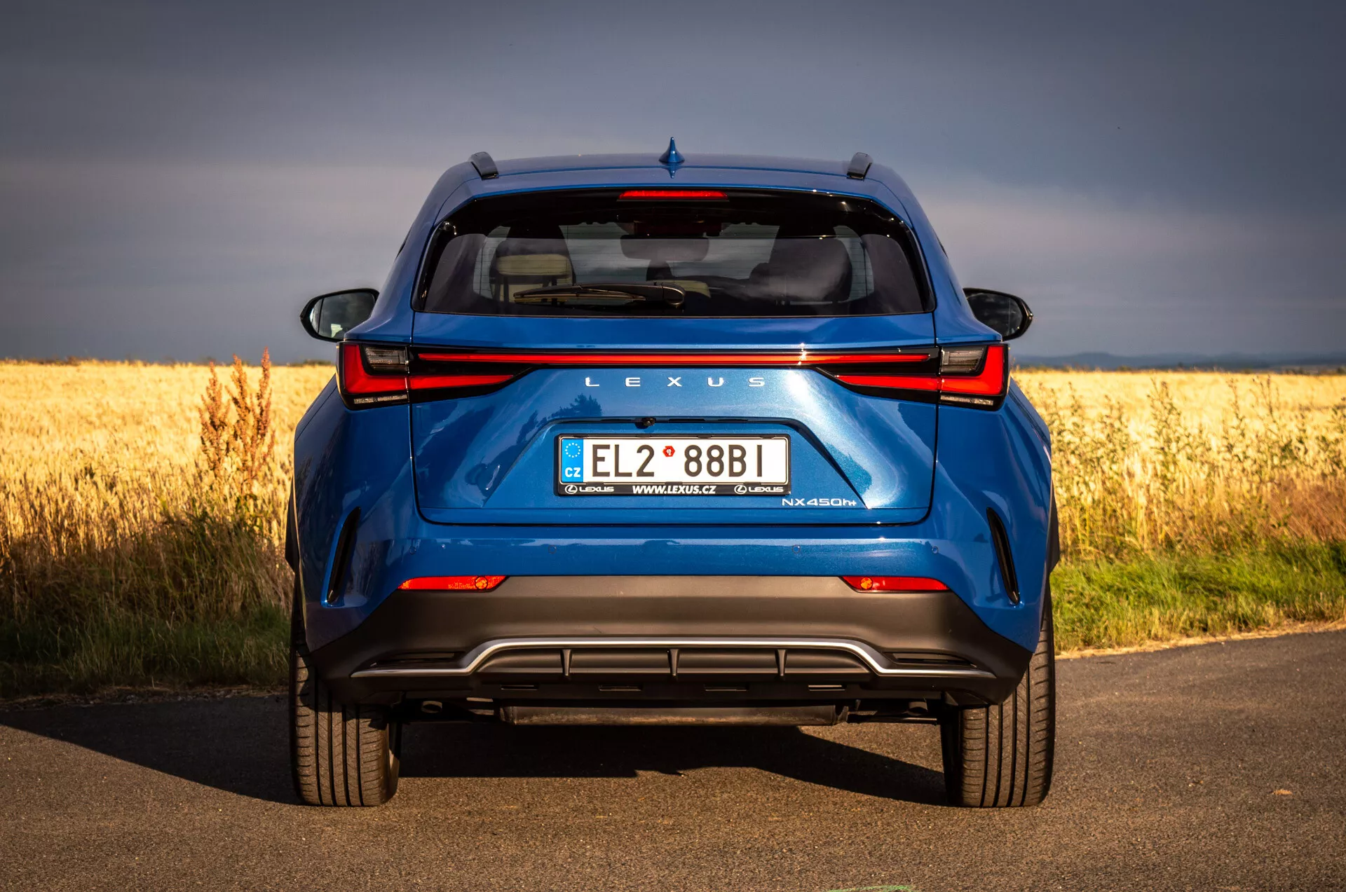 Zadný pohľad na modré SUV Lexus NX 2021 na asfaltovej ceste so zlatým poľom v pozadí, zdôrazňujúci hybridnú technológiu a dizajnové prvky