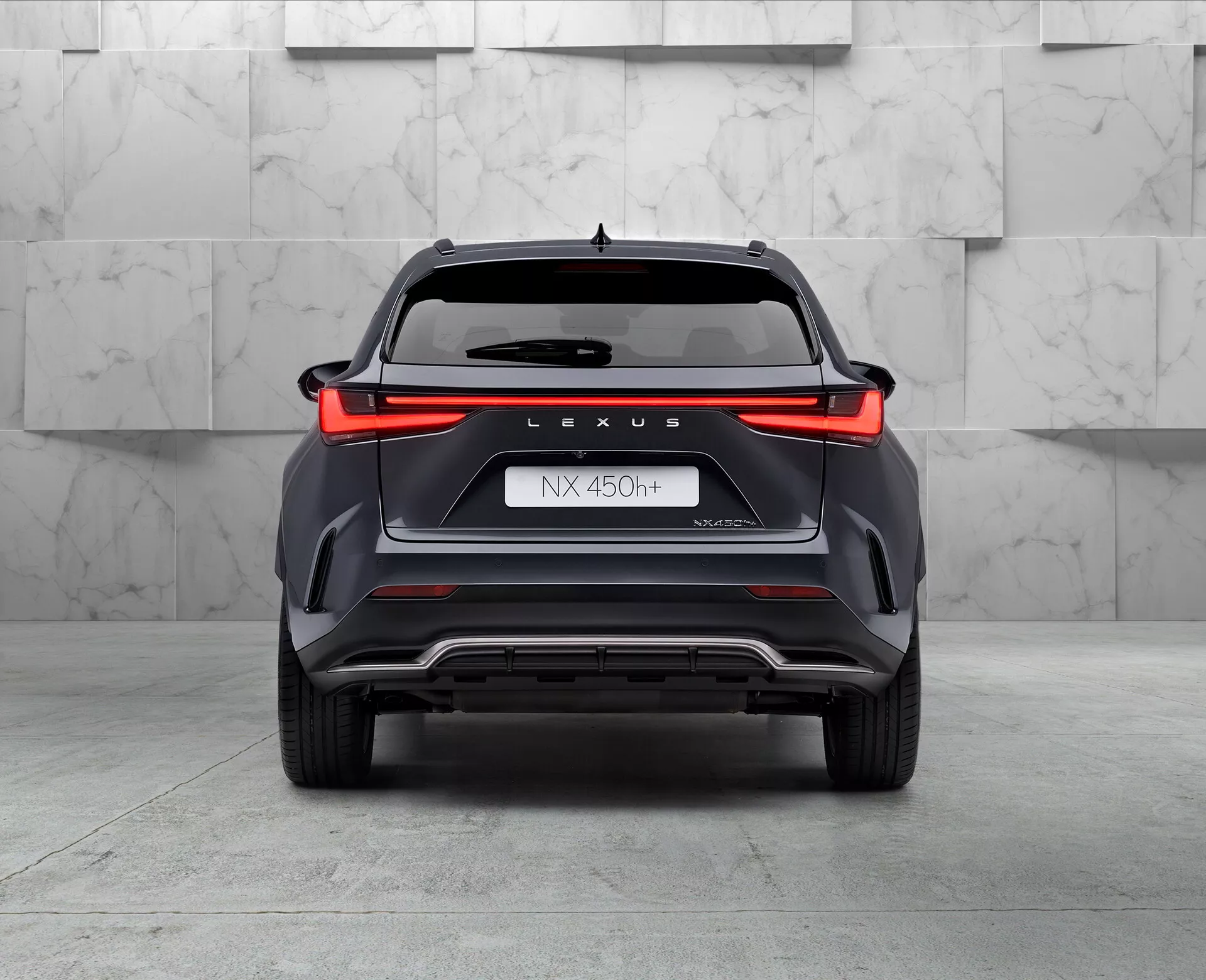 Zadný pohľad na hybridné SUV Lexus NX 450h, zachytený z nízkeho uhla mierne nakloneného doľava, ktorý zobrazuje dizajn svetiel po celej šírke, značku 'LEXUS', označenie modelu 'NX 450h', názov 'NEXUS' a symbol hybridu 'h'. Nárazník s uhlovými štýlovými prvkami, ventilačnými vlastnosťami a strieborným ochranným plechom, s viditeľným dvojitým výfukovým hrotom na ľavej strane, v prostredí showroomu s vyleštenou podlahou a neutrálnym pozadím.