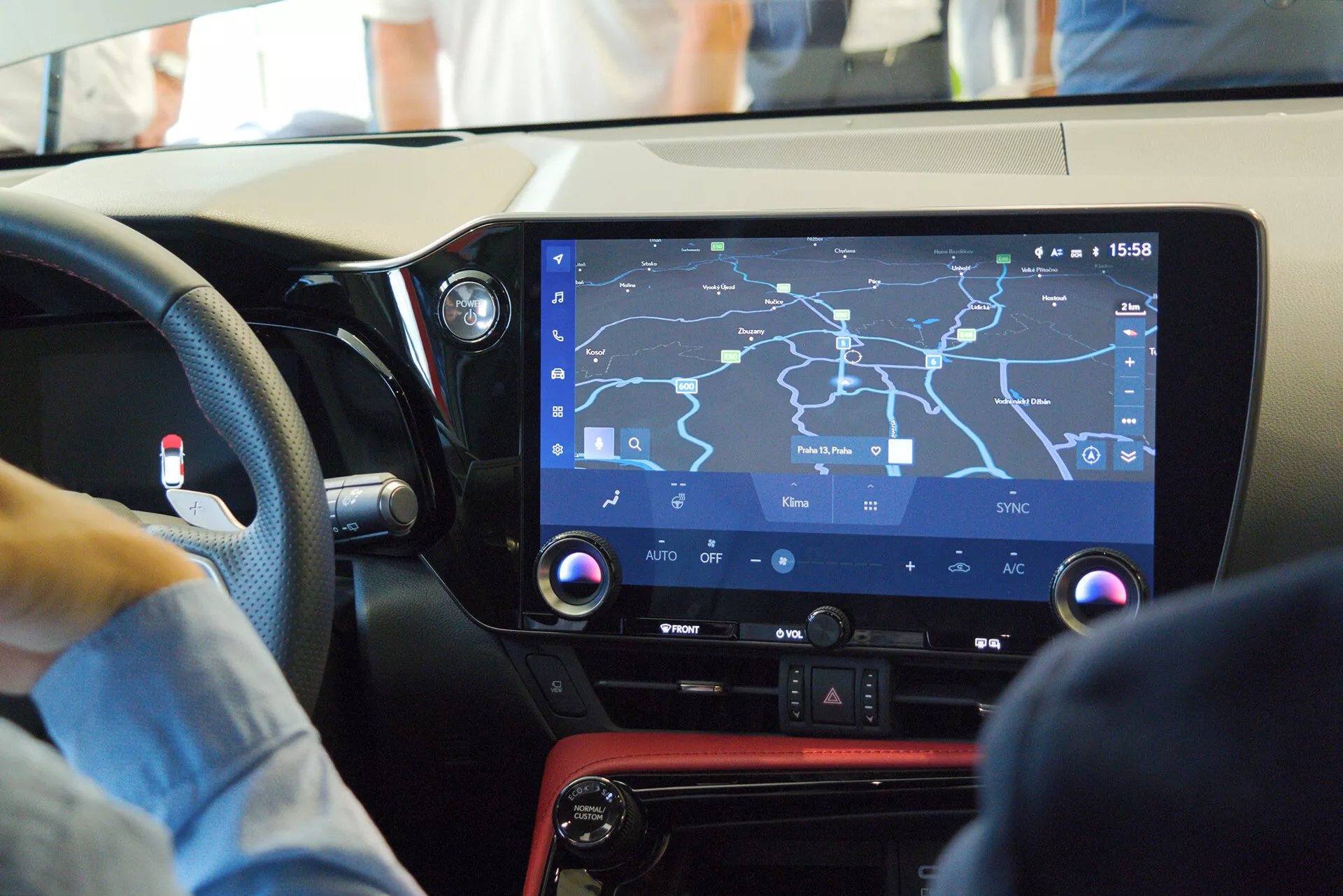 Pohľad na interiér moderného auta zameraný na centrálnu konzolu a infotainment systém z pohľadu spolujazdca, zobrazujúci veľký dotykový displej s navigačnou mapou, fyzické ovládacie prvky klimatických funkcií, lesklý čierny panel s červeným akcentom a naznačujúci vodičsky prívetivý dizajn v rušnom prostredí.