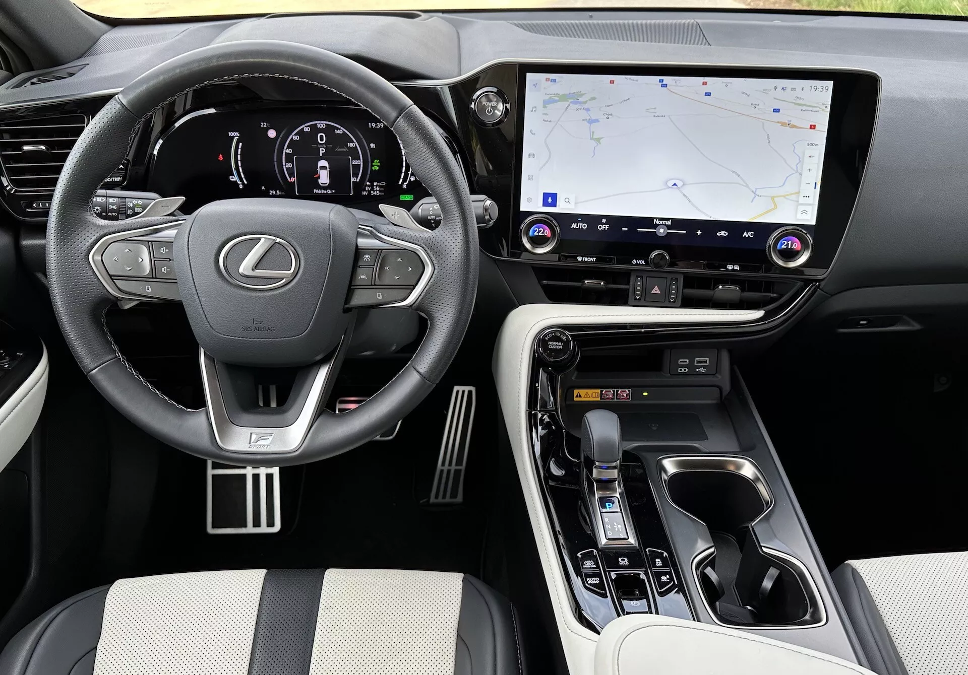 Interiérový pohľad na Lexus NX 2021 zobrazujúci oblasť vodiča a stredovú konzolu zvýšeného uhla, s logom Lexus na volante s namontovanými ovládačmi, osvetleným digitálnym prístrojovým panelom, veľkým dotykovým informačno-zábavným systémom s GPS navigáciou, tlačidlami pre ovládanie klímy a zvuku, ergonomickým voličom prevodov, ovládačmi režimu jazdy, luxusnými materiálmi na sedadlách a dotykových bodoch, kovovými akcentmi a viditeľnými držiakmi na nápoje a úložným priestorom.
