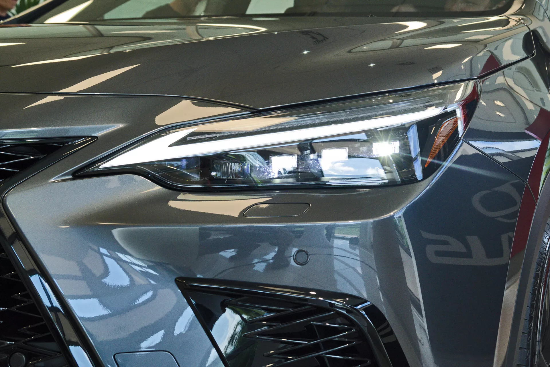 Detailný pohľad na prednú časť modelu Lexus NX 2021, zameraný na svetlomet na strane vodiča s pokročilou LED osvetľovacou technológiou a časť agresívnej mriežky. Obrázok zachytáva odraz lesklého kovového strieborného laku a dynamické línie, ktoré prispievajú k aerodynamickej estetike vozidla. Fotografované v interiéri, pravdepodobne v showroome, zdôrazňujúce luxusný dizajn a vysokokvalitnú povrchovú úpravu vozidla.
