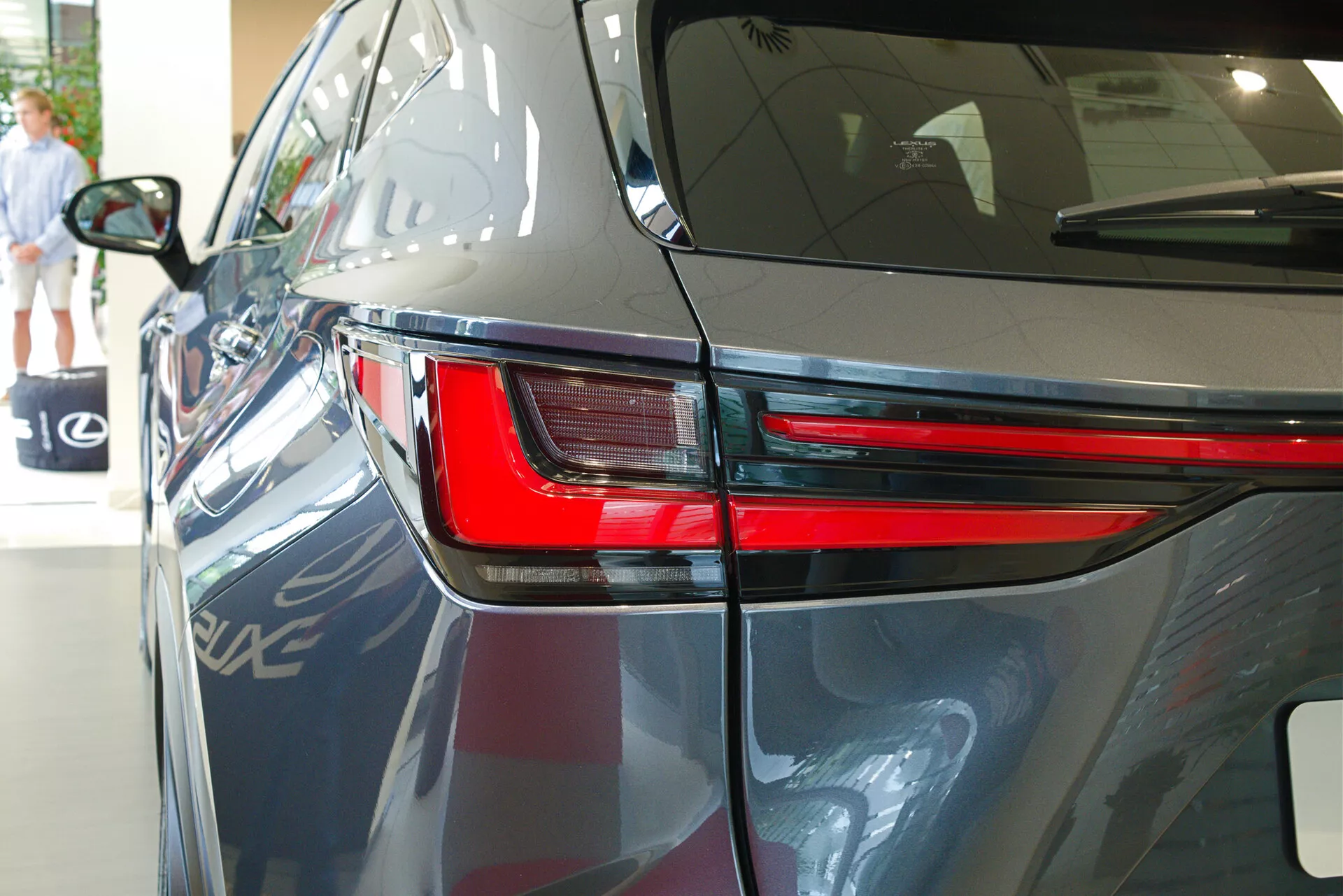 Blízky pohľad na Lexus NX 2021 zozadu v troch štvrtinách uhla, ktorý zdôrazňuje elegantný dizajn LED zadného svetla, časť zadného štvrtpanelu a časť veka kufra. Obrázok ukazuje lesklý tmavý lak auta, aerodynamické línie karosérie a naznačuje vnútorný výstavný priestor s odrazmi ďalšieho vozidla, architektonickými prvkami a osobou kráčajúcou v pozadí.
